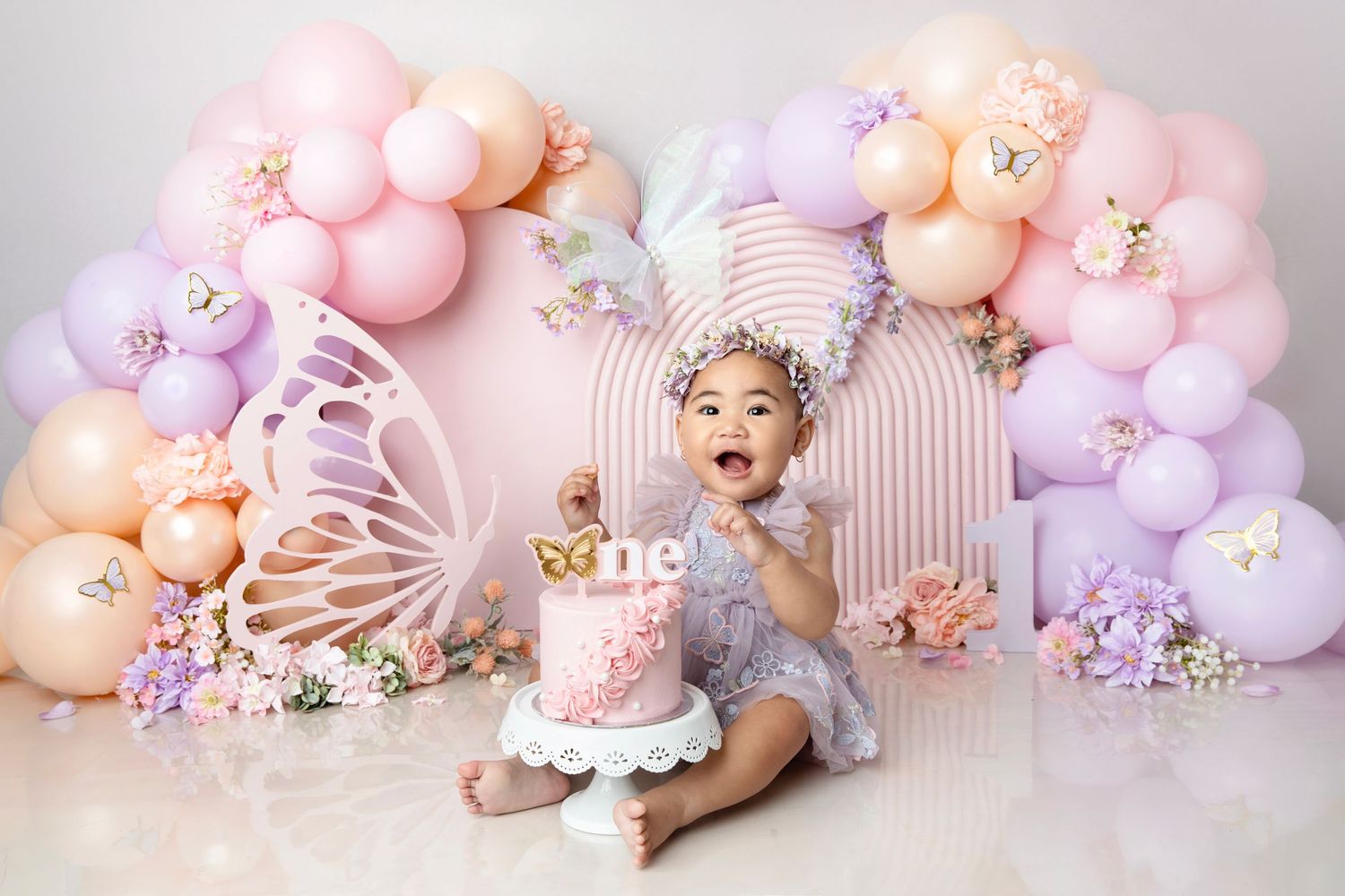 Baby girl in gray dress enjoys first birthday cake smash with pastel balloon arch and butterfly decorations.