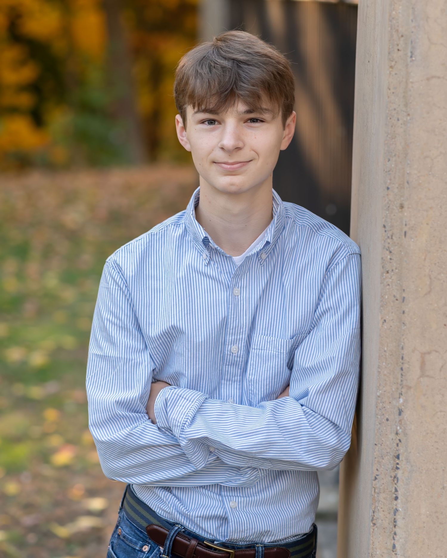 Young man in light blue striped shirt leaning against stone pillar with arms crossed, smiling outdoors in autumn.