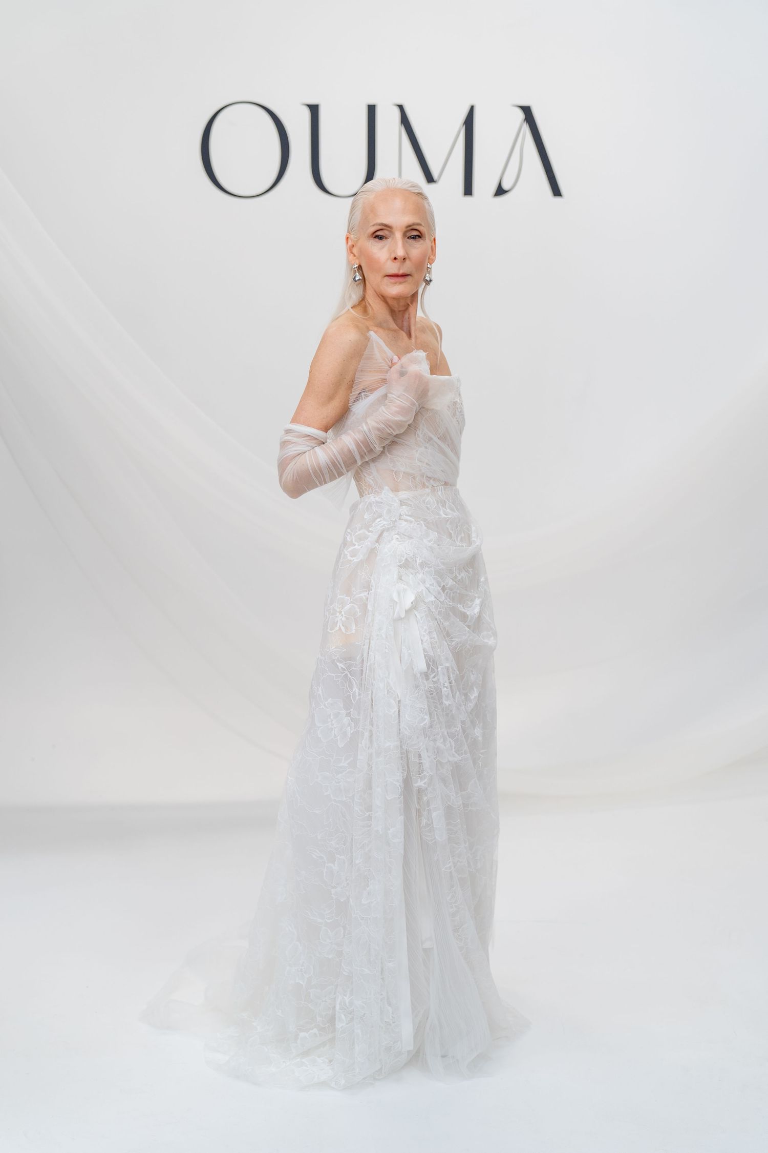 Elegant older woman in white lace bridal gown with sheer gloves posing before OUMA backdrop.