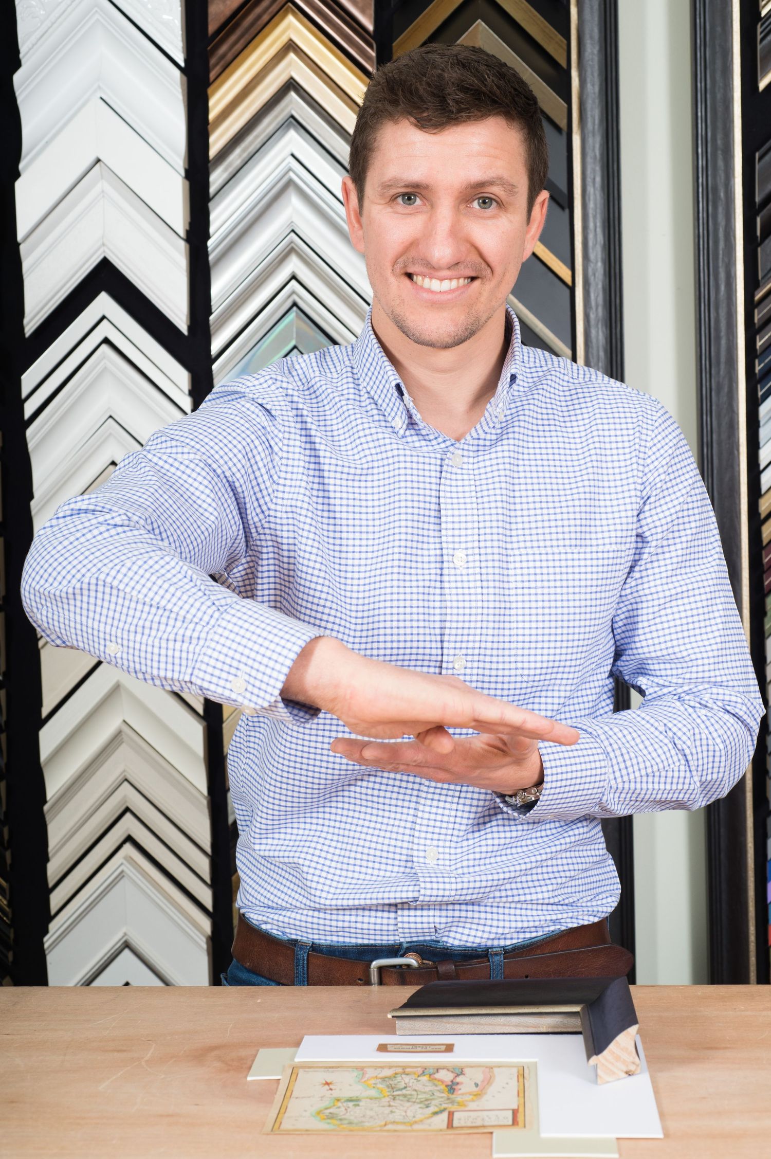 Smiling man in a checkered shirt demonstrating framing with his hands. Background has various picture frame samples. Table holds maps and fr