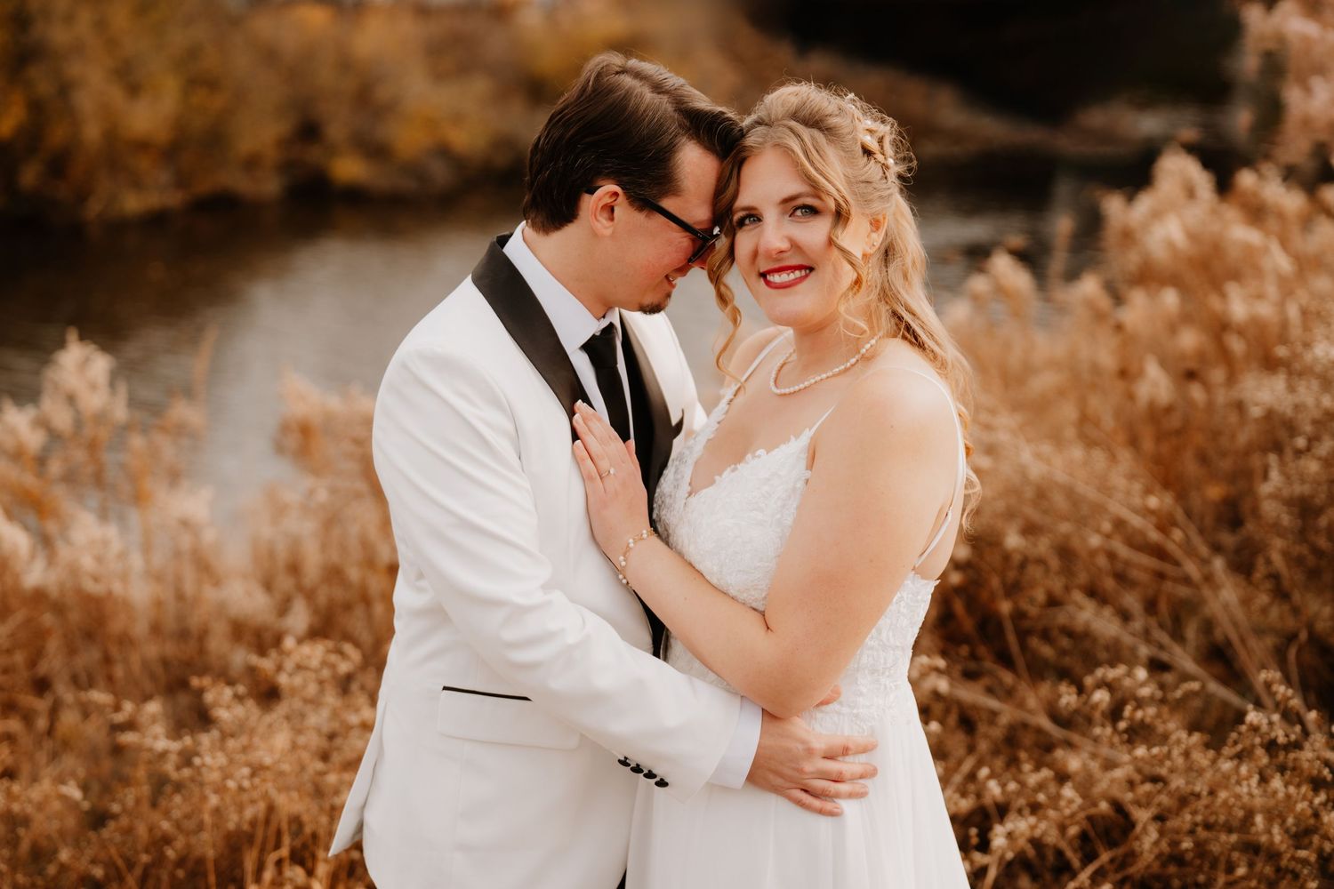A bride and groom embrace outdoors in autumn, surrounded by golden tall grasses near a serene pond.