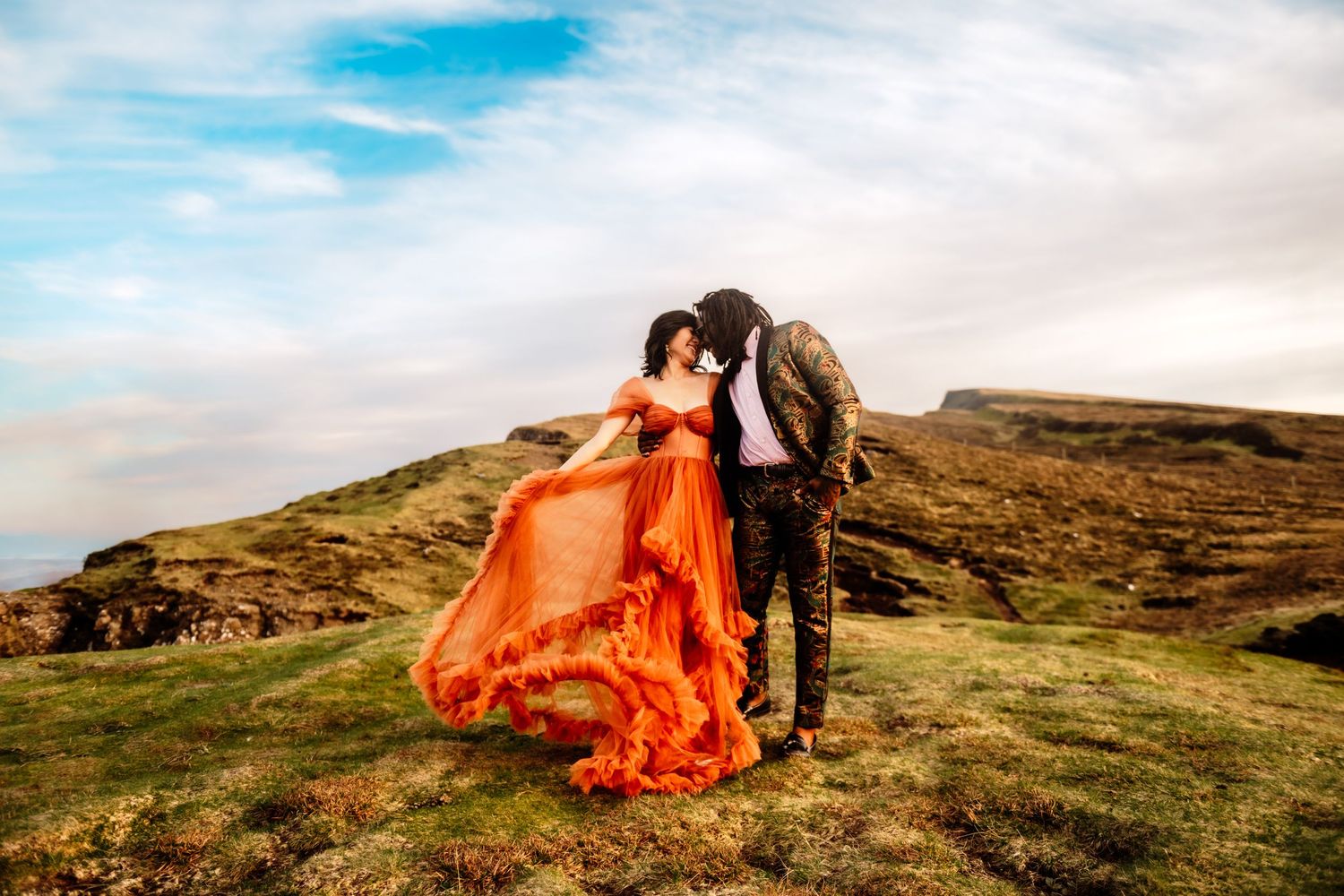 Couple in stunning orange gown embracing on scenic highland landscape with dramatic cloudy sky backdrop.