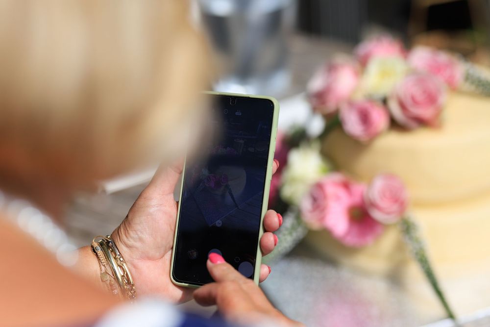 Woman with pink nails photographing a wedding cake decorated with pink roses using a smartphone.