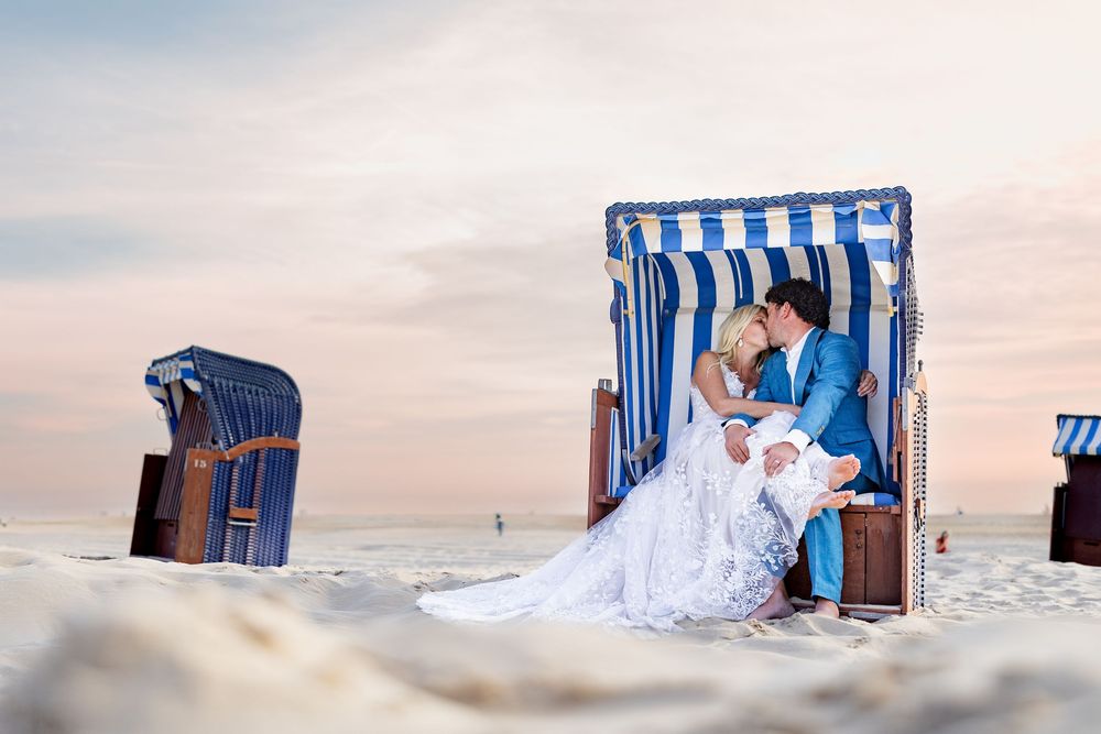Bride and groom share a romantic kiss in a blue striped beach chair on a sandy shore at sunset.