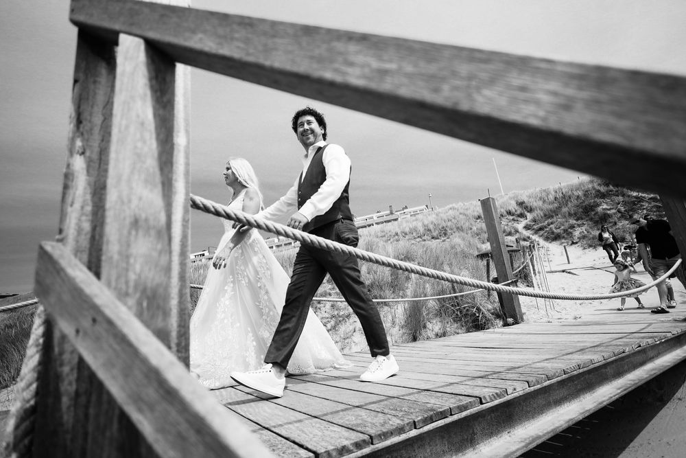 Black and white photo of a bride and groom walking on a wooden bridge with rope railings on their wedding day.