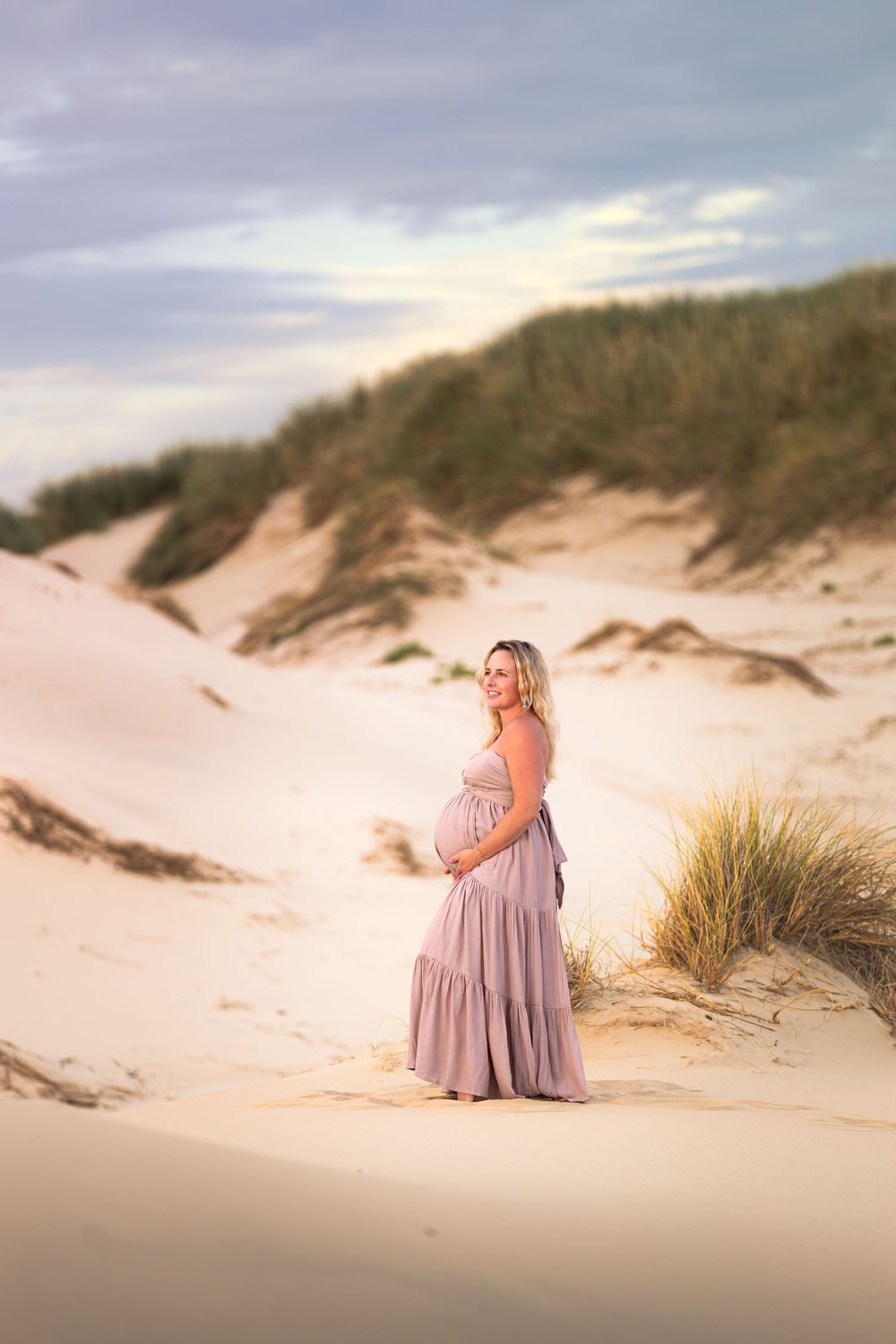 Pregnant woman in mauve dress cradling baby bump on sandy dunes at golden hour.