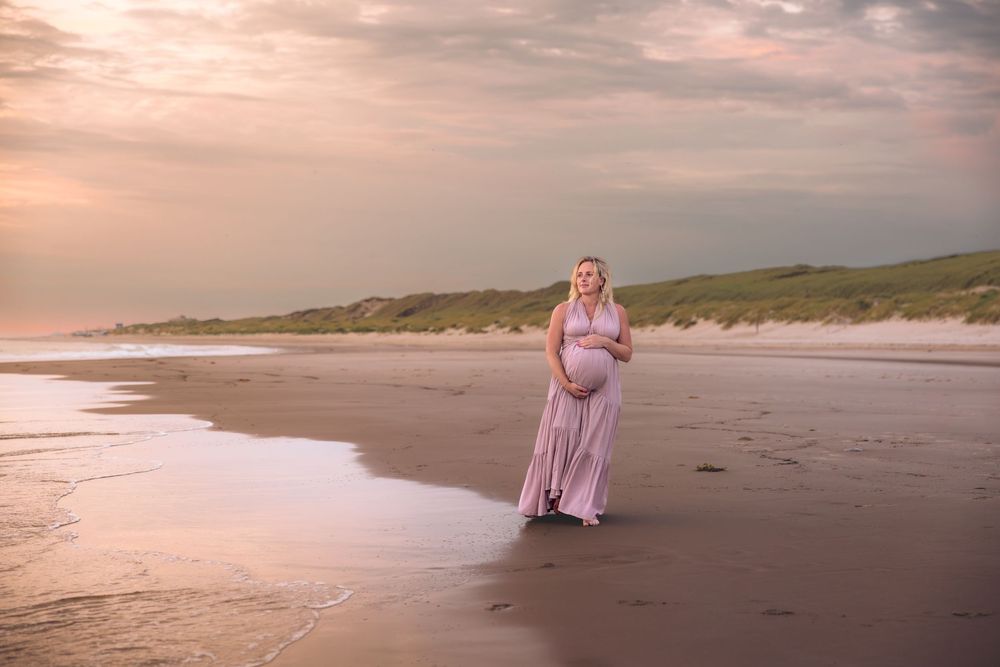 Pregnant woman in pink gown standing on beach at golden sunset with sand dunes in background.
