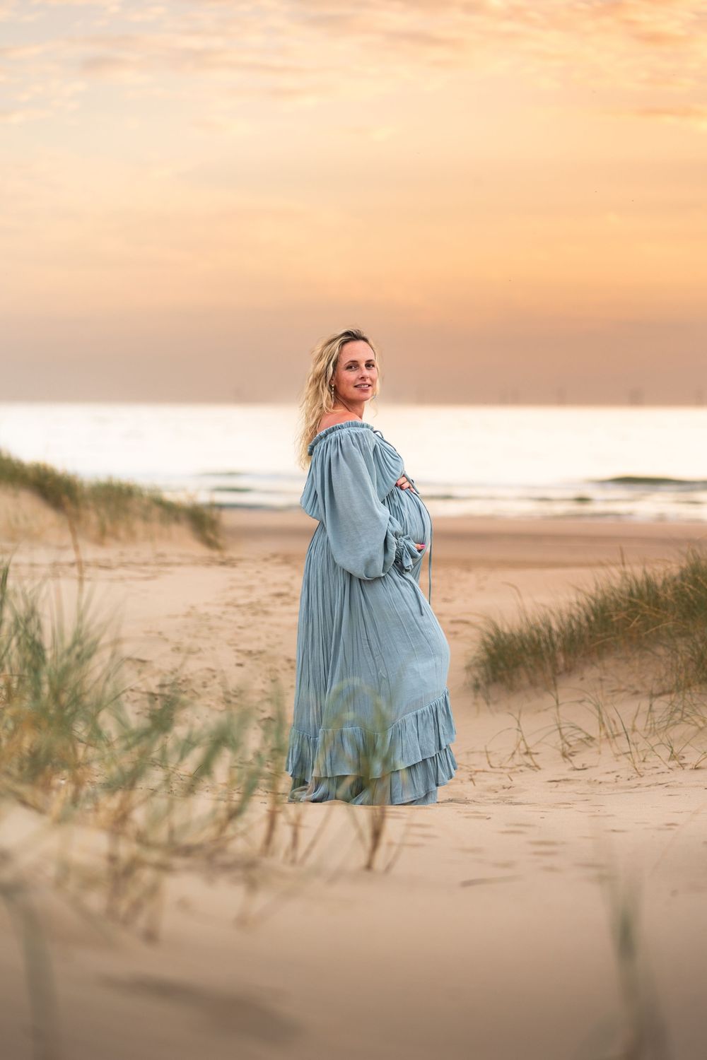 Pregnant woman in blue dress standing on sandy beach dunes at sunset, smiling over shoulder.
