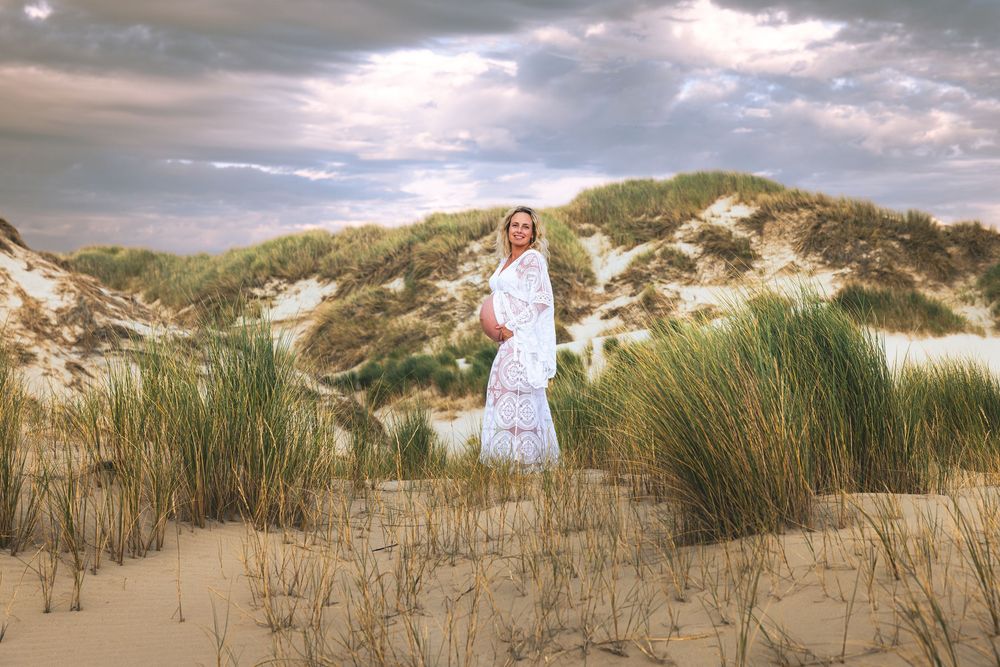 Pregnant woman in white lace dress standing among sand dunes with green grass under dramatic cloudy sky.