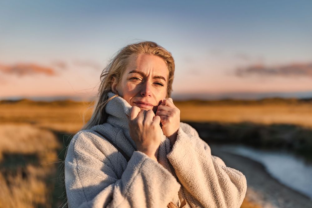 Blonde woman in cozy white fleece jacket holding collar near face outdoors at golden sunset hour.