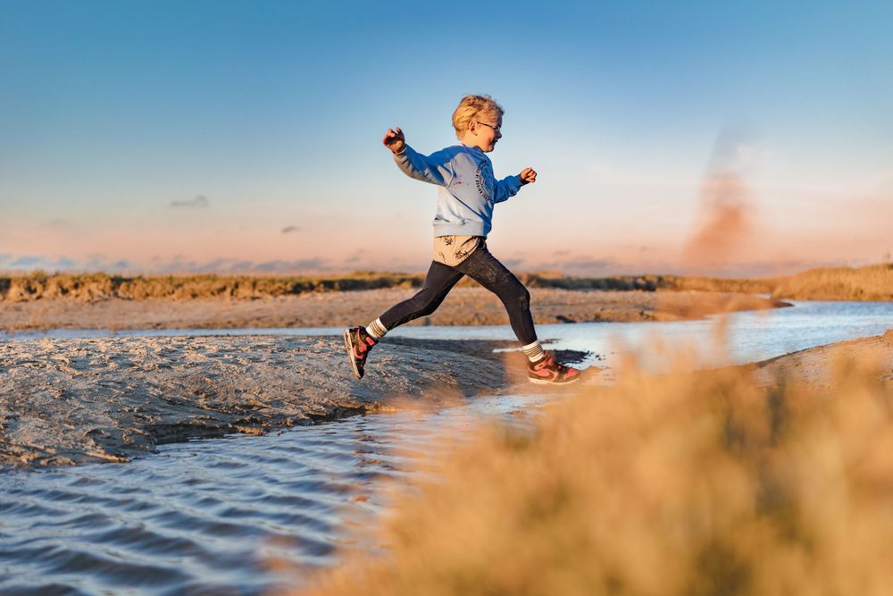 Child joyfully leaping over shallow water on a sandy beach at sunset, wearing casual athletic clothing.