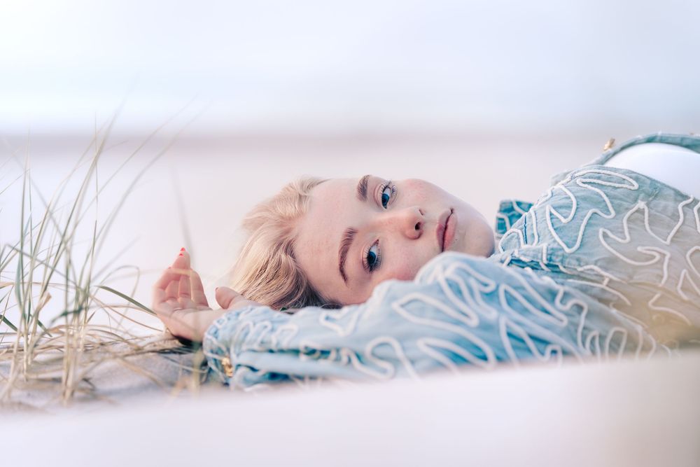 A young blonde child with blue eyes lying on sandy ground outdoors, wearing a light blue patterned outfit.