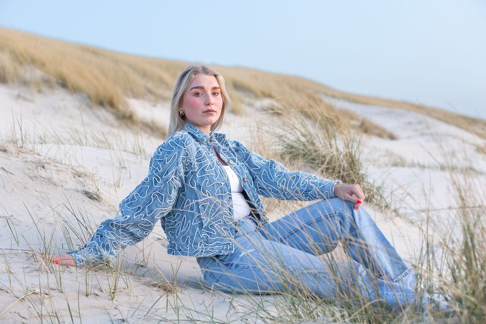 Young woman in blue patterned denim jacket and jeans sitting on sandy beach dunes with grass.