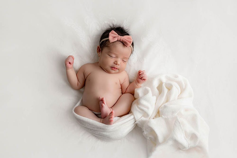 Sleeping newborn baby girl in white wrap with pink bow headband on white fluffy background by professional Temecula newborn photographer.