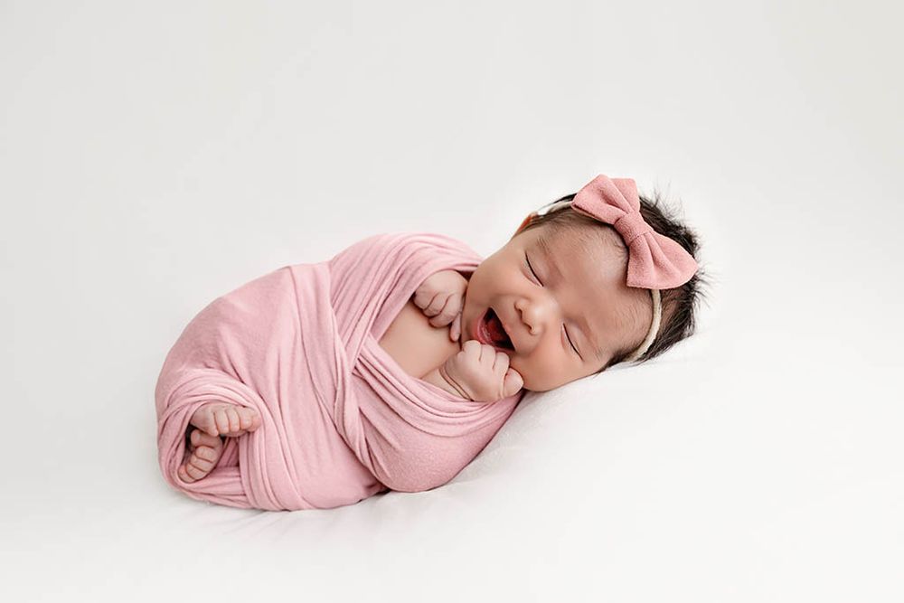 Affordable Temecula newborn photographer - Smiling newborn baby girl swaddled in pink wrap with bow headband on white background.