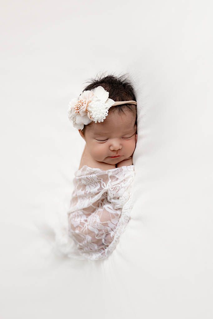 Temecula newborn photographer photo of sleeping baby girl wrapped in white lace swaddle with floral headband on white background.