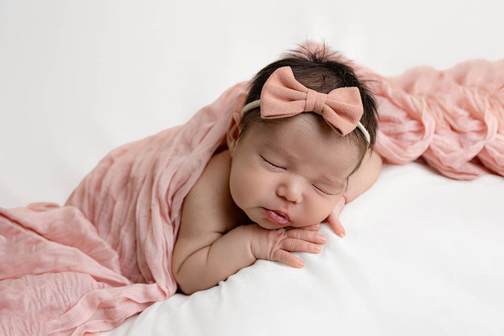 Sleeping newborn baby girl wrapped in a pink blanket wearing a pink bow headband on a white background.