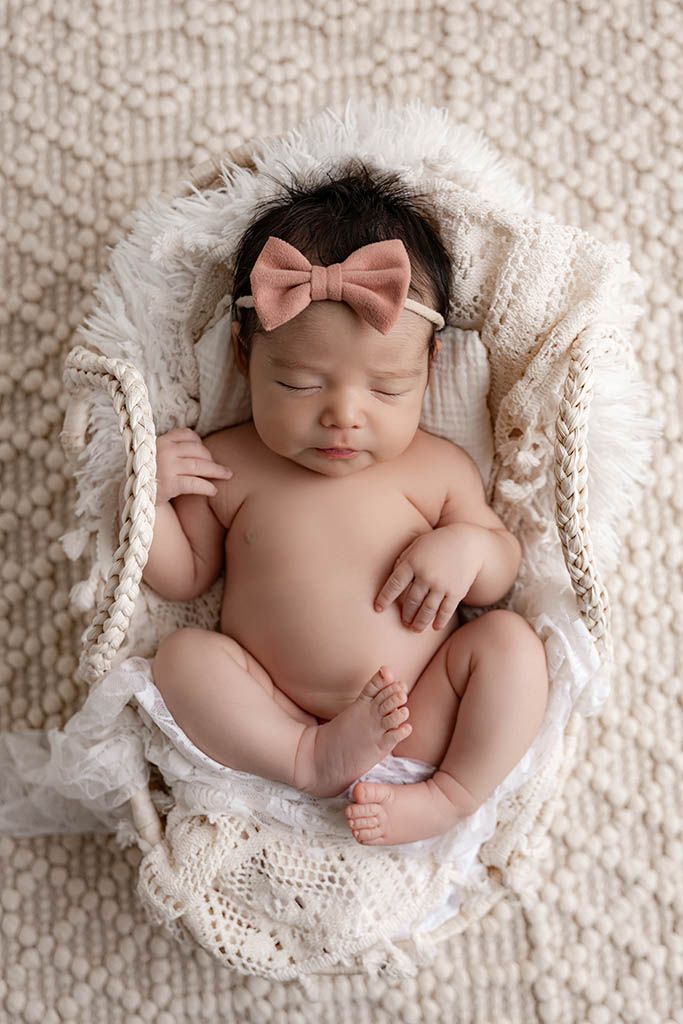Temecula newborn photography - baby girl in a woven basket wearing a pink bow headband, surrounded by soft cream blankets.