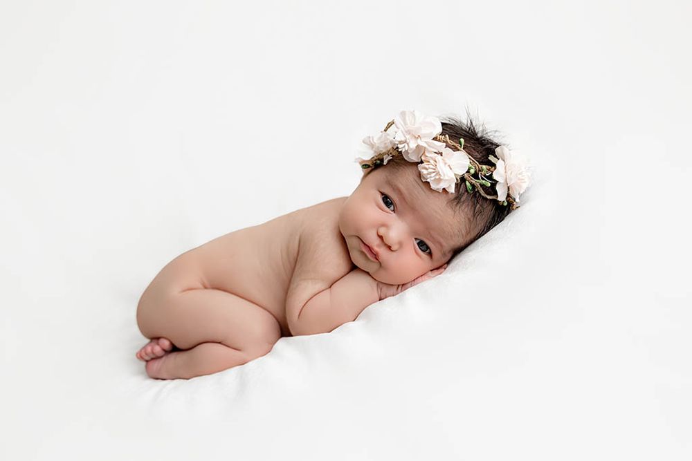 Newborn baby girl lying on white blanket wearing a delicate pink floral crown, peaceful studio portrait by Temecula newborn photographer.