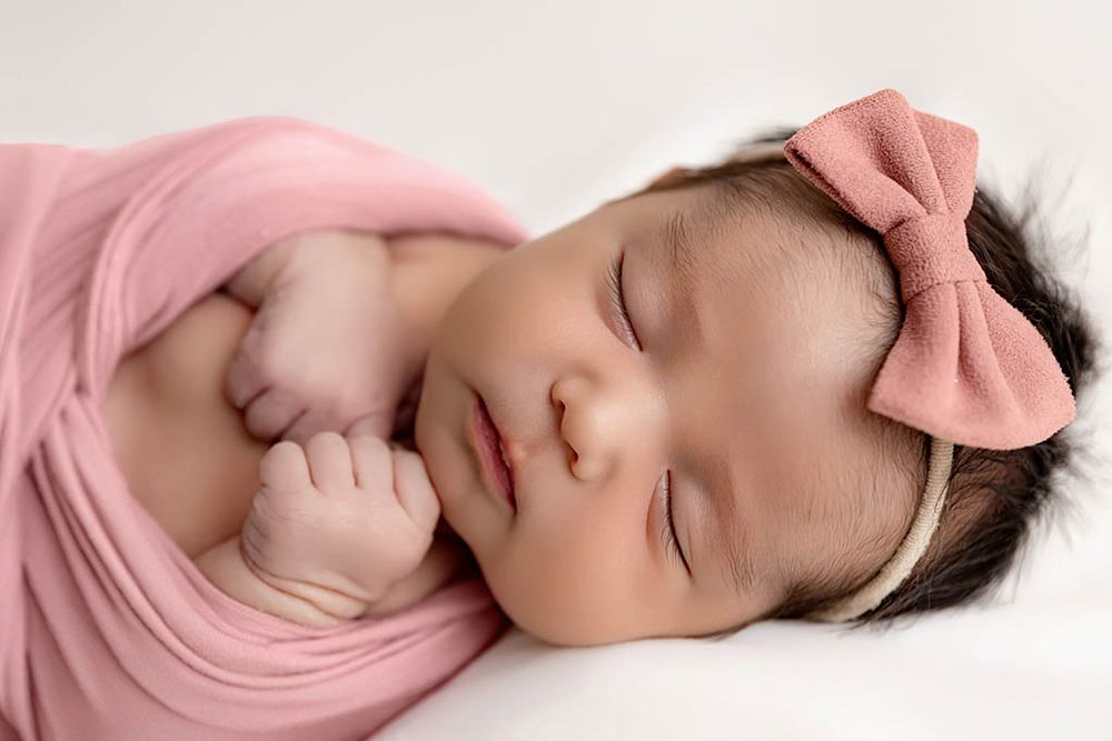 Peaceful newborn baby girl sleeping wrapped in pink swaddle with bow headband on white background by Temecula newborn photographer.
