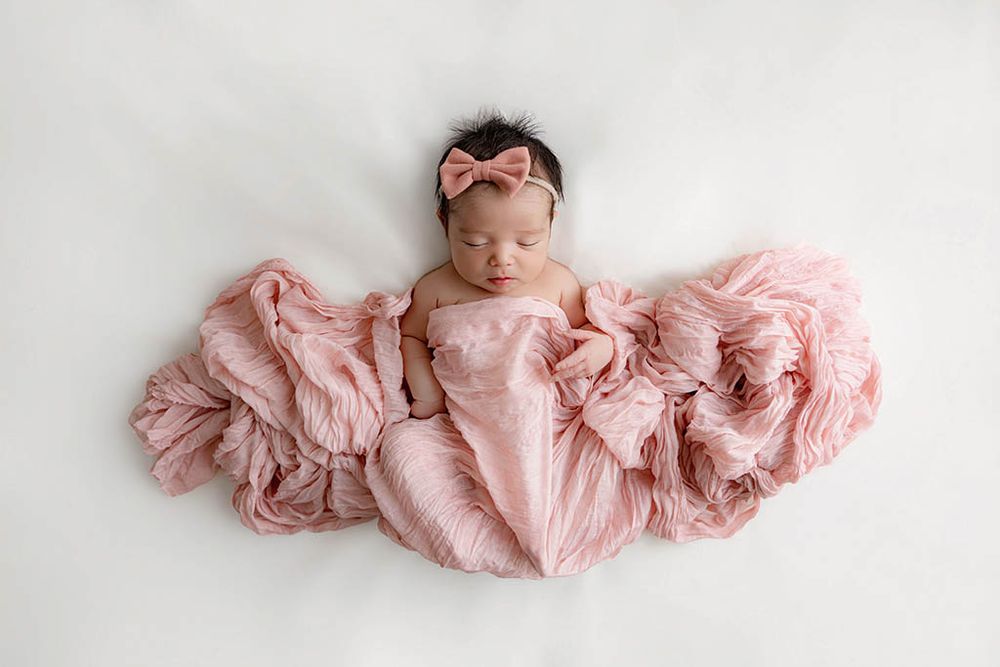 Timeless newborn photography in Temecula CA - Sleeping newborn baby girl wrapped in pink fabric with a bow headband on white background.