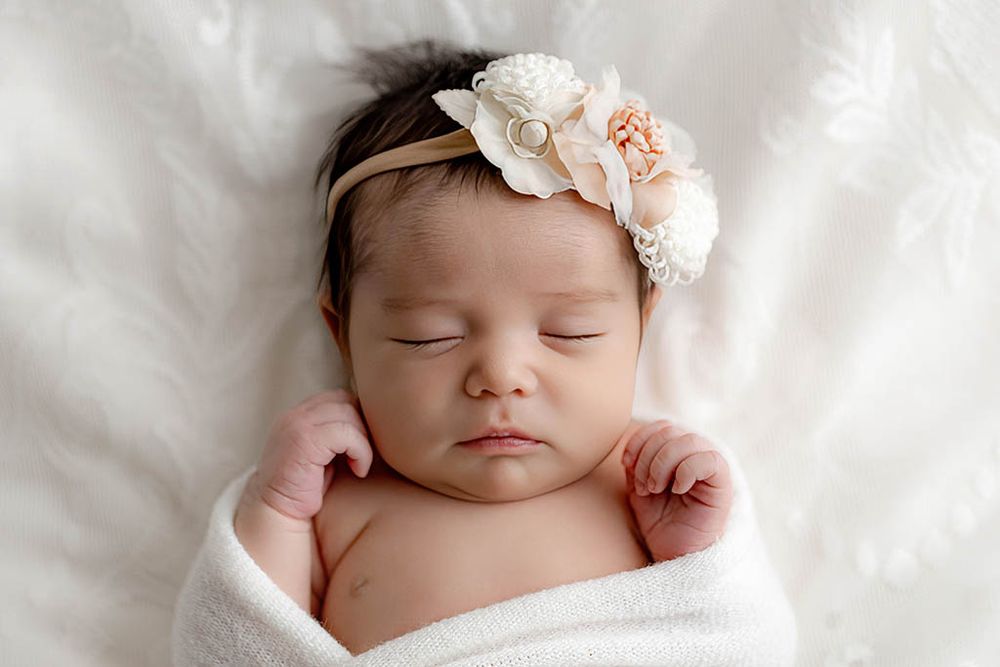 Sleeping newborn baby girl wrapped in white swaddle with floral headband on soft white background.