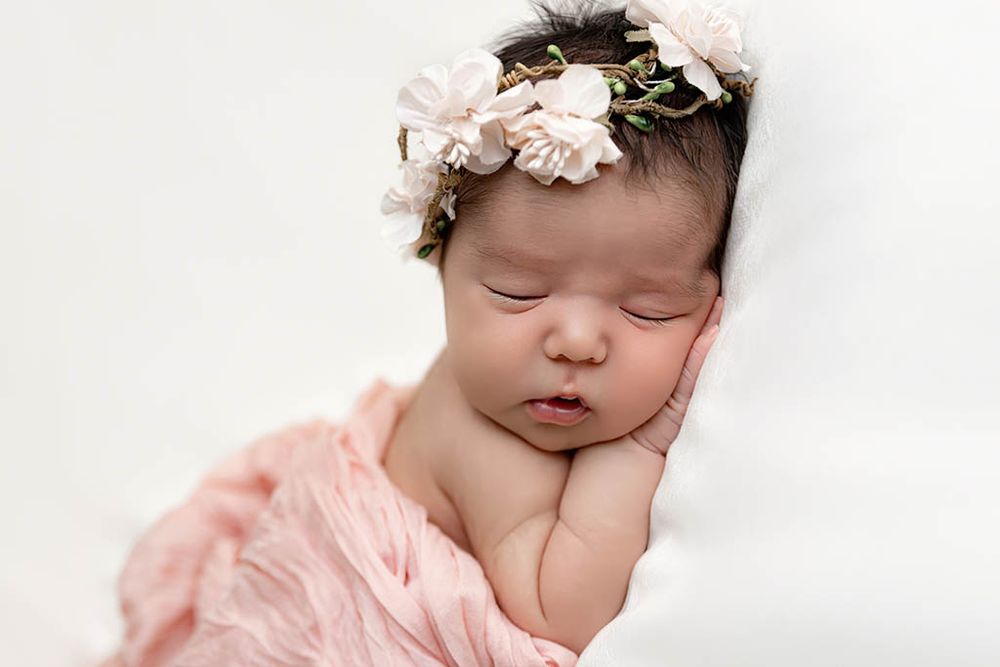 Temecula newborn photographer photo of peaceful newborn baby girl in pink wrap with floral crown headband sleeping on white backdrop.