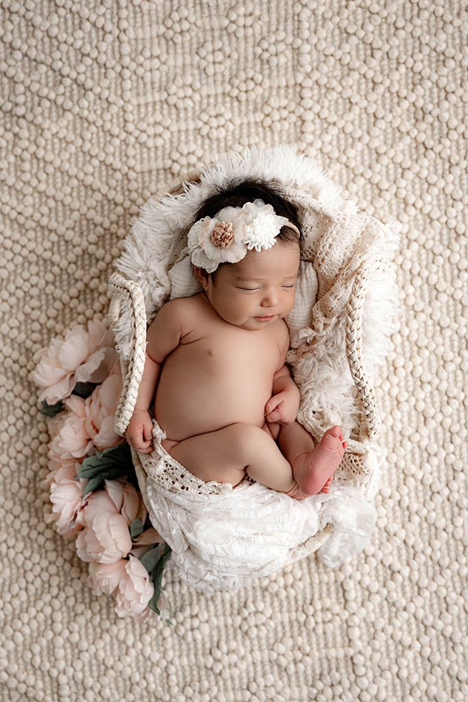 Professional Temecula newborn photography studio photo of baby girl sleeping in a wicker basket with pink peonies.