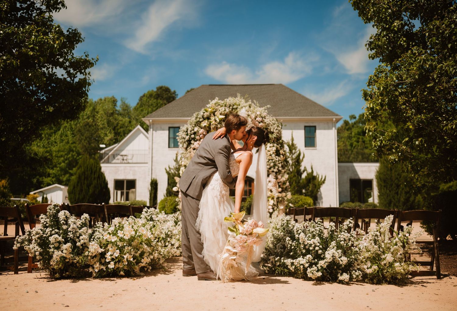 Bride and groom sharing a kiss under a floral arch with white blooms outside an elegant white wedding venue.