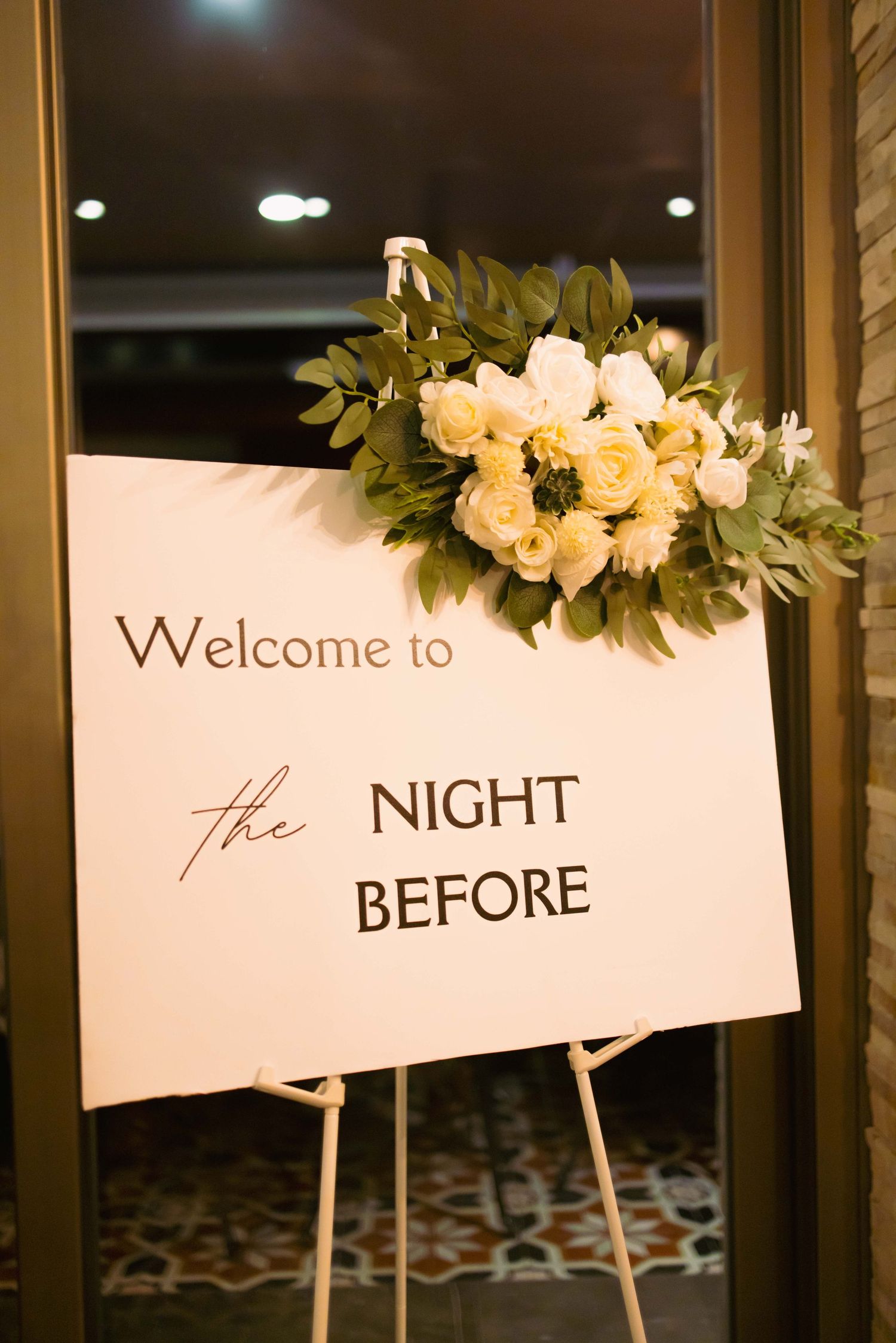 Elegant wedding rehearsal dinner welcome sign with white roses and greenery on a white easel stand.