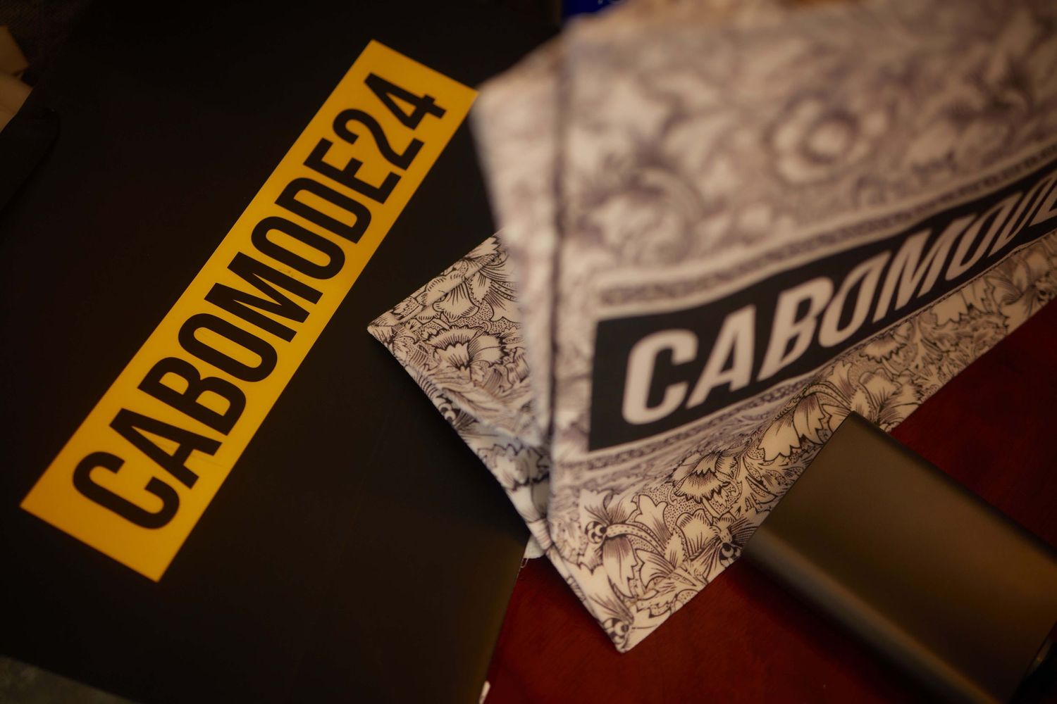 Two CABOMODE24 branded items, a yellow sticker and a patterned bandana, displayed on a dark surface.