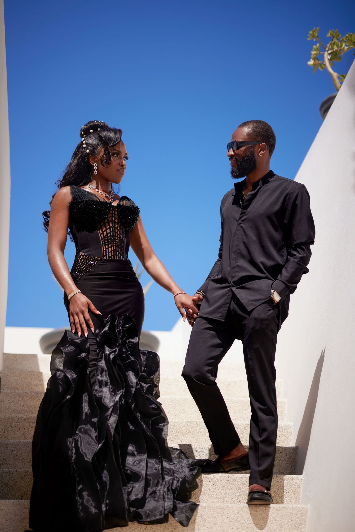 Elegant Black couple in black formal attire holding hands on white outdoor staircase under blue sky.