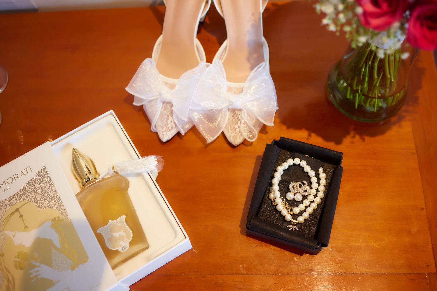 White bridal heels with bow detail beside pearl necklace in box, perfume bottle, and pink roses on wooden table.