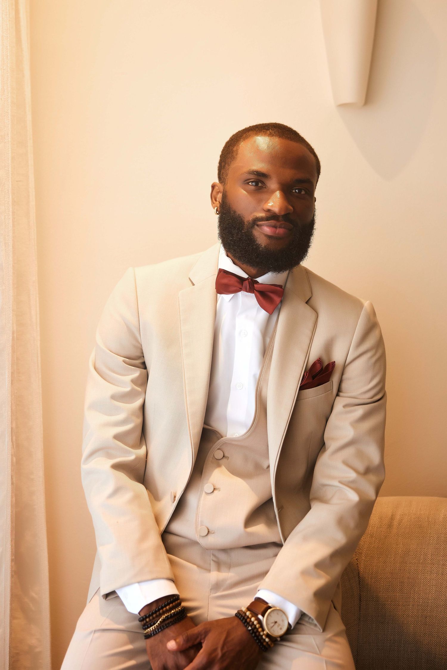 A well-dressed Black man in a beige three-piece suit with a burgundy bow tie and pocket square sits confidently.