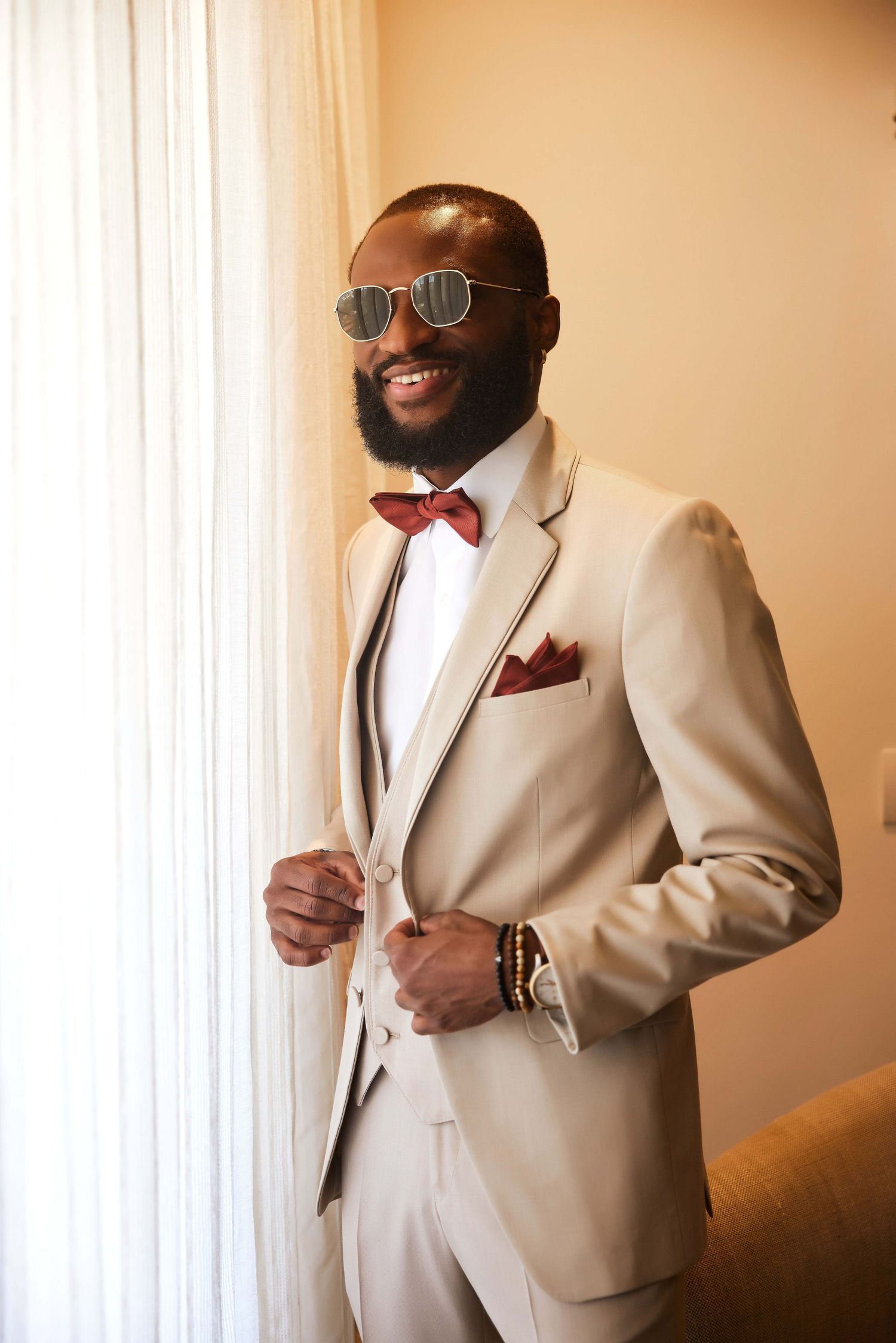 Smiling man in beige suit, red bow tie, pocket square, sunglasses, and bracelets posing confidently near a window.