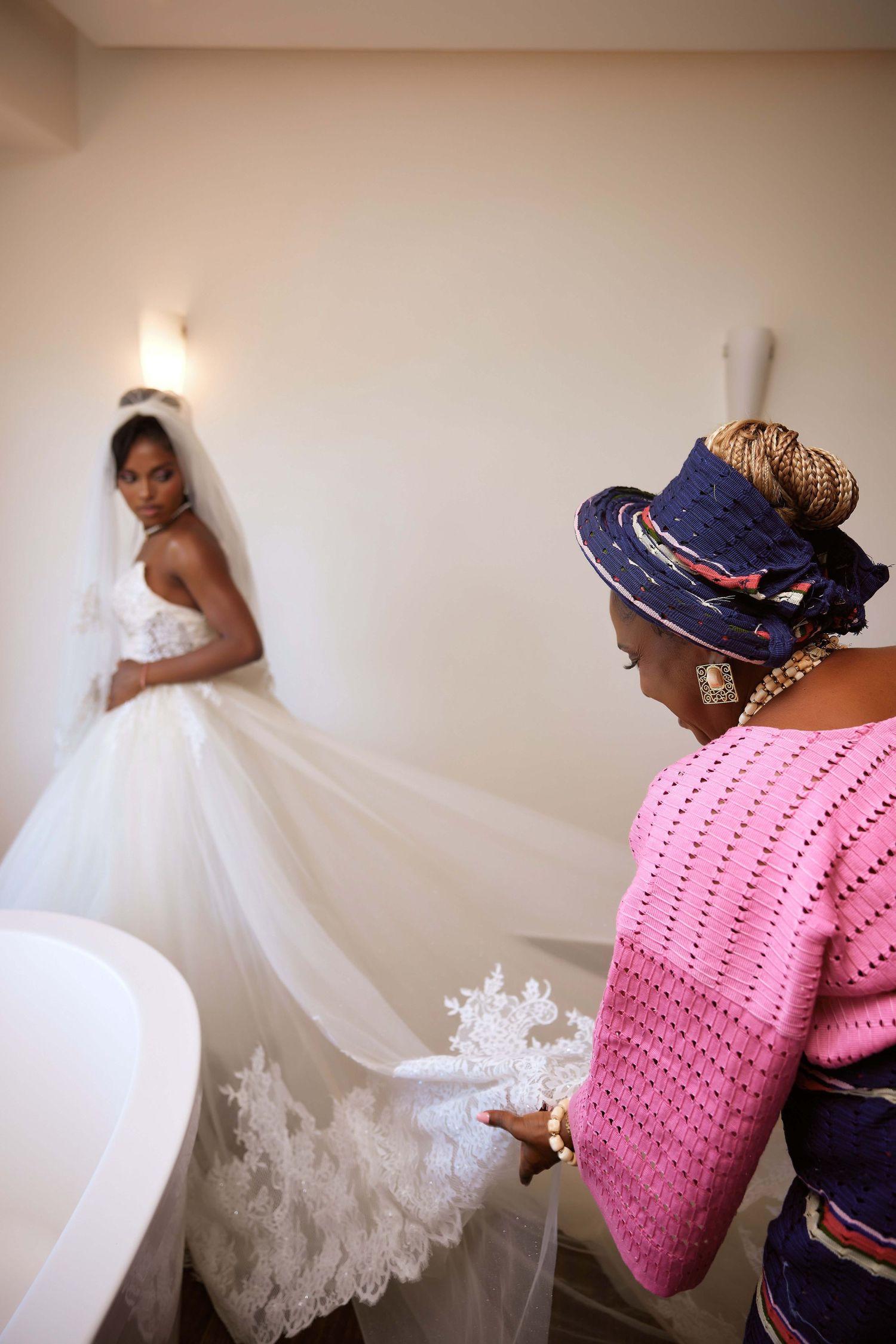 A bride in a white gown and veil stands as a woman in pink adjusts her lace train during wedding preparations.