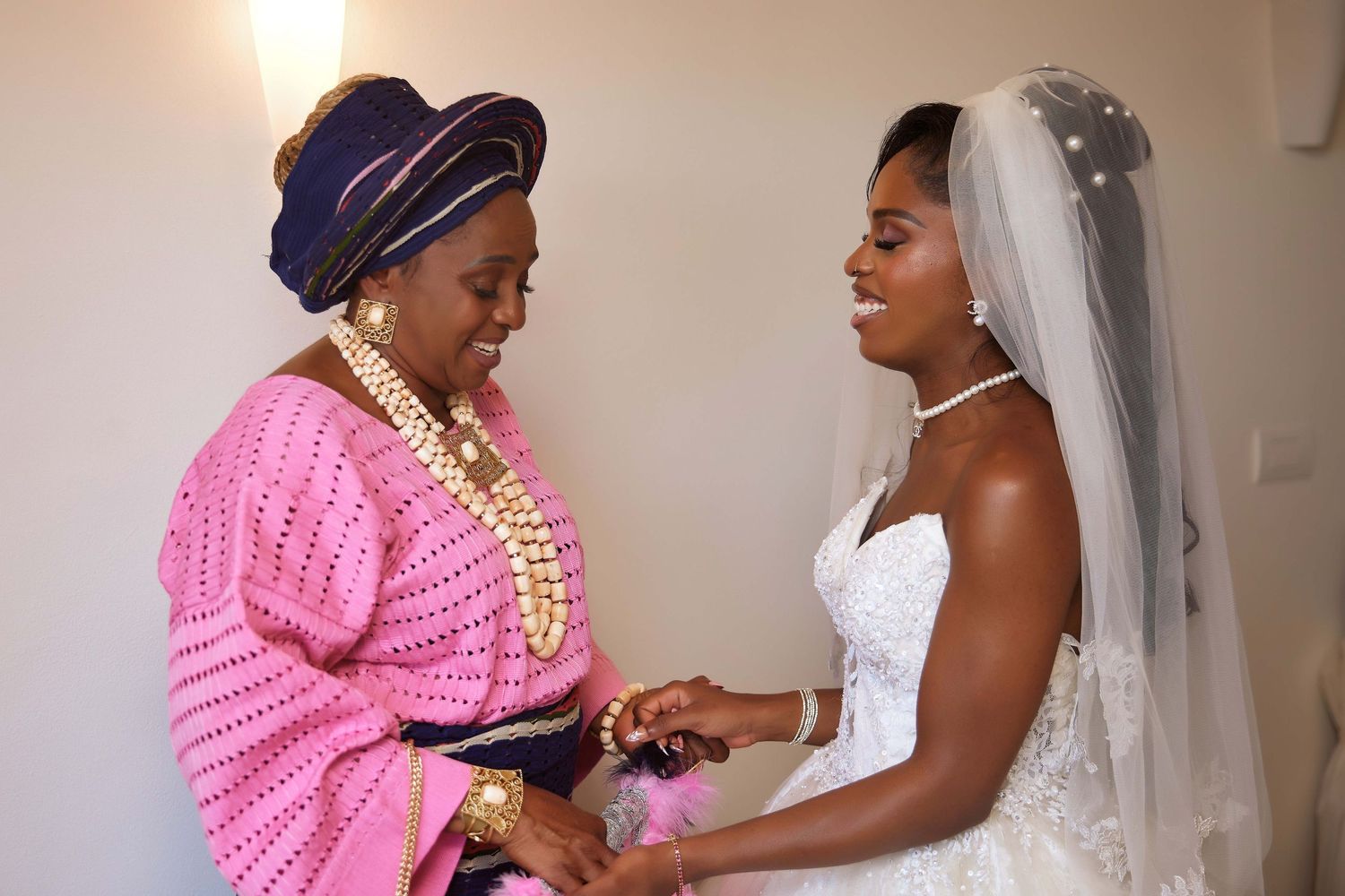 A bride in a white wedding dress and veil holds hands with an older woman in a pink outfit and gold jewelry.