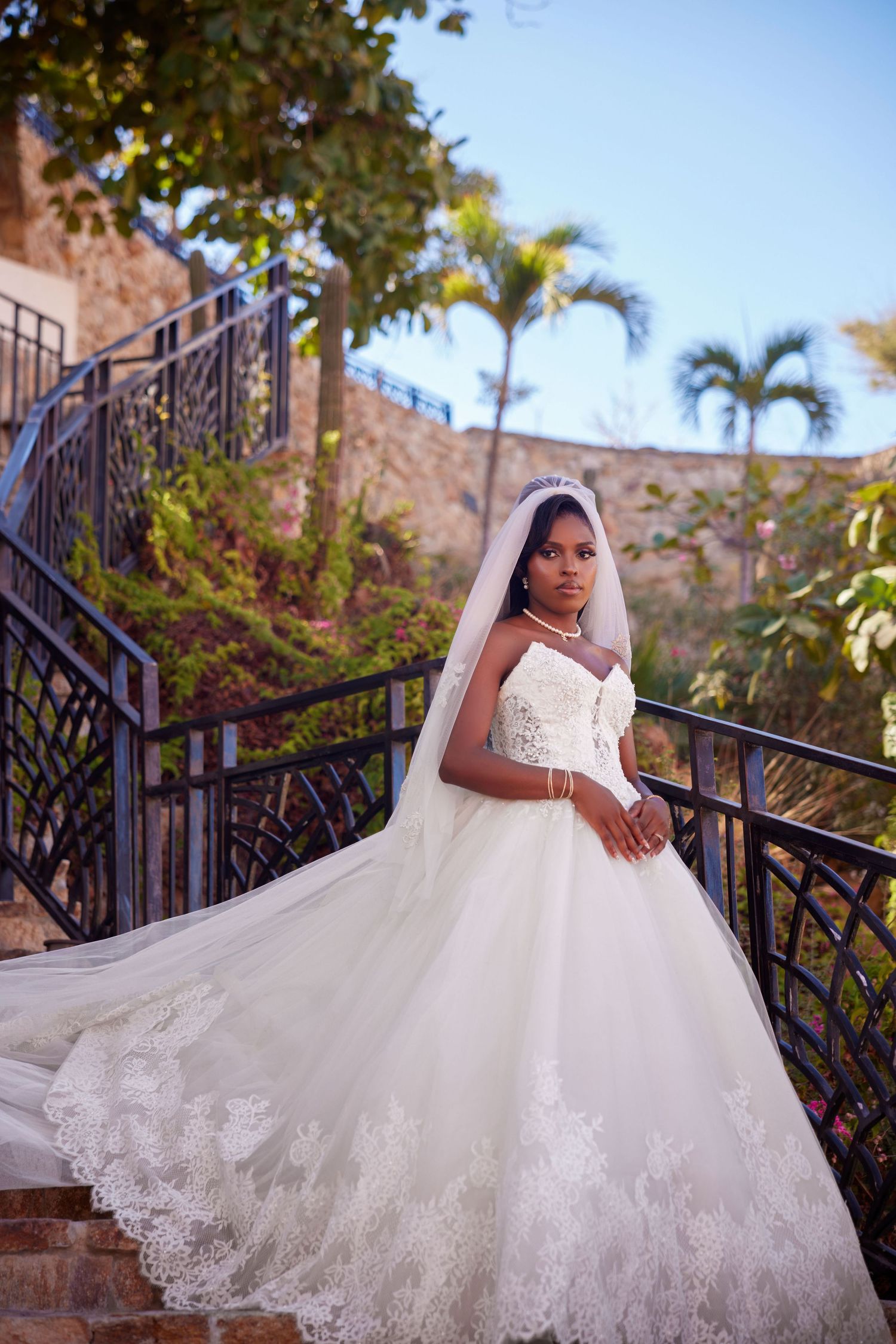 A bride in a white lace ball gown and veil poses gracefully on outdoor stairs surrounded by tropical greenery.