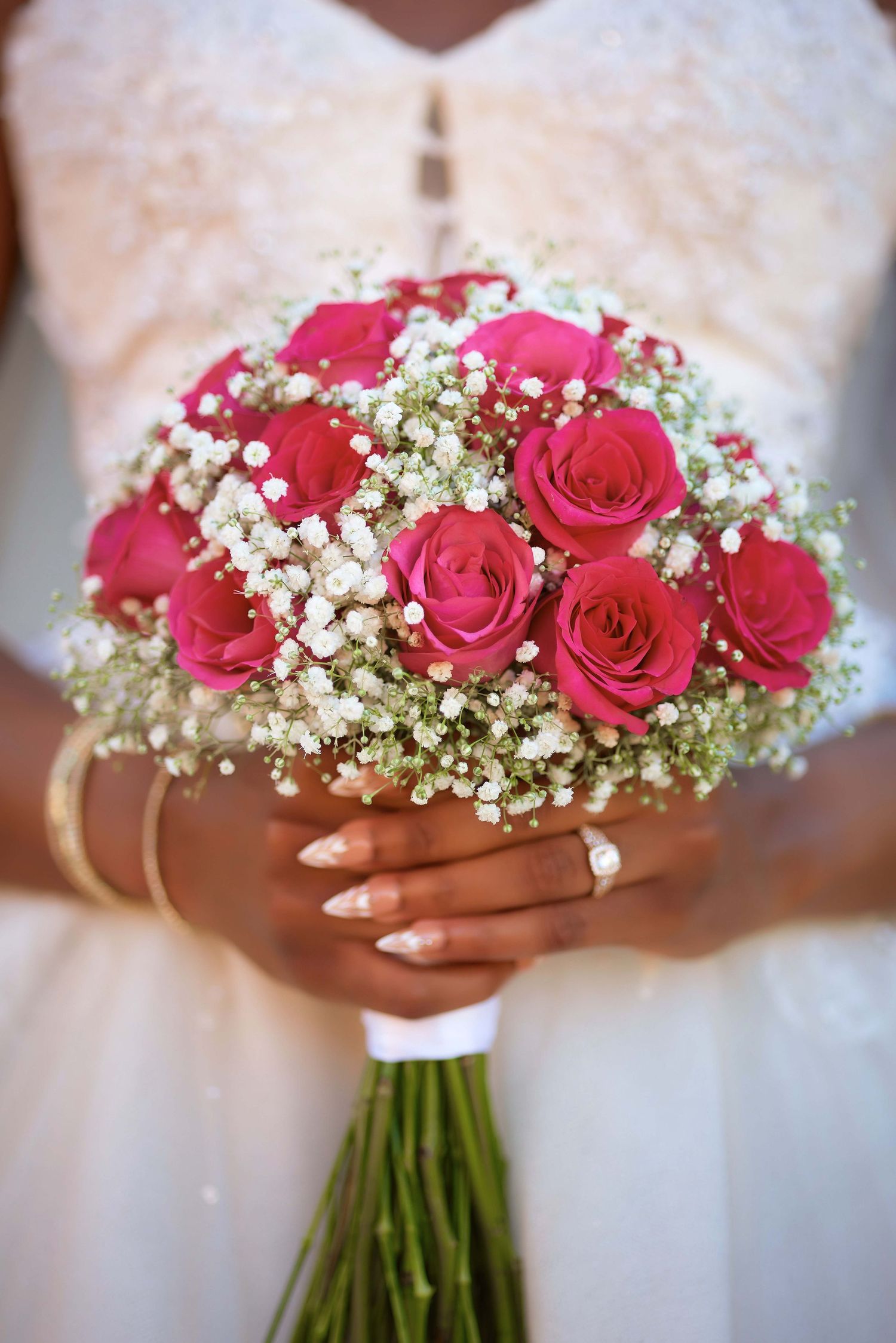Bride in white dress holding a bridal bouquet of pink roses and white baby's breath with gold bracelet and ring.