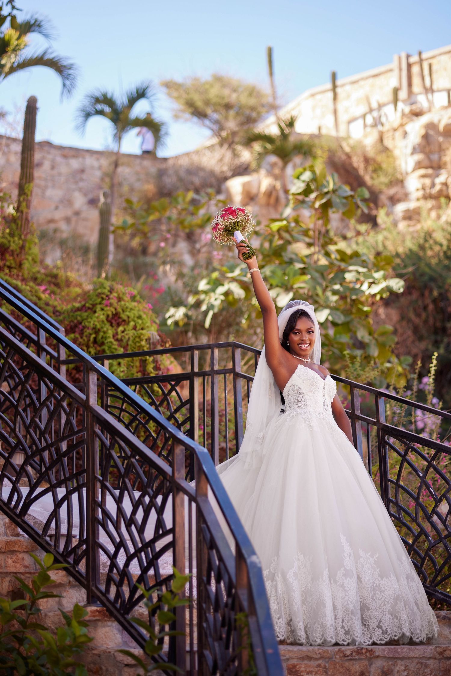 Joyful Black bride in white ball gown raises bouquet on ornate staircase surrounded by tropical gardens.