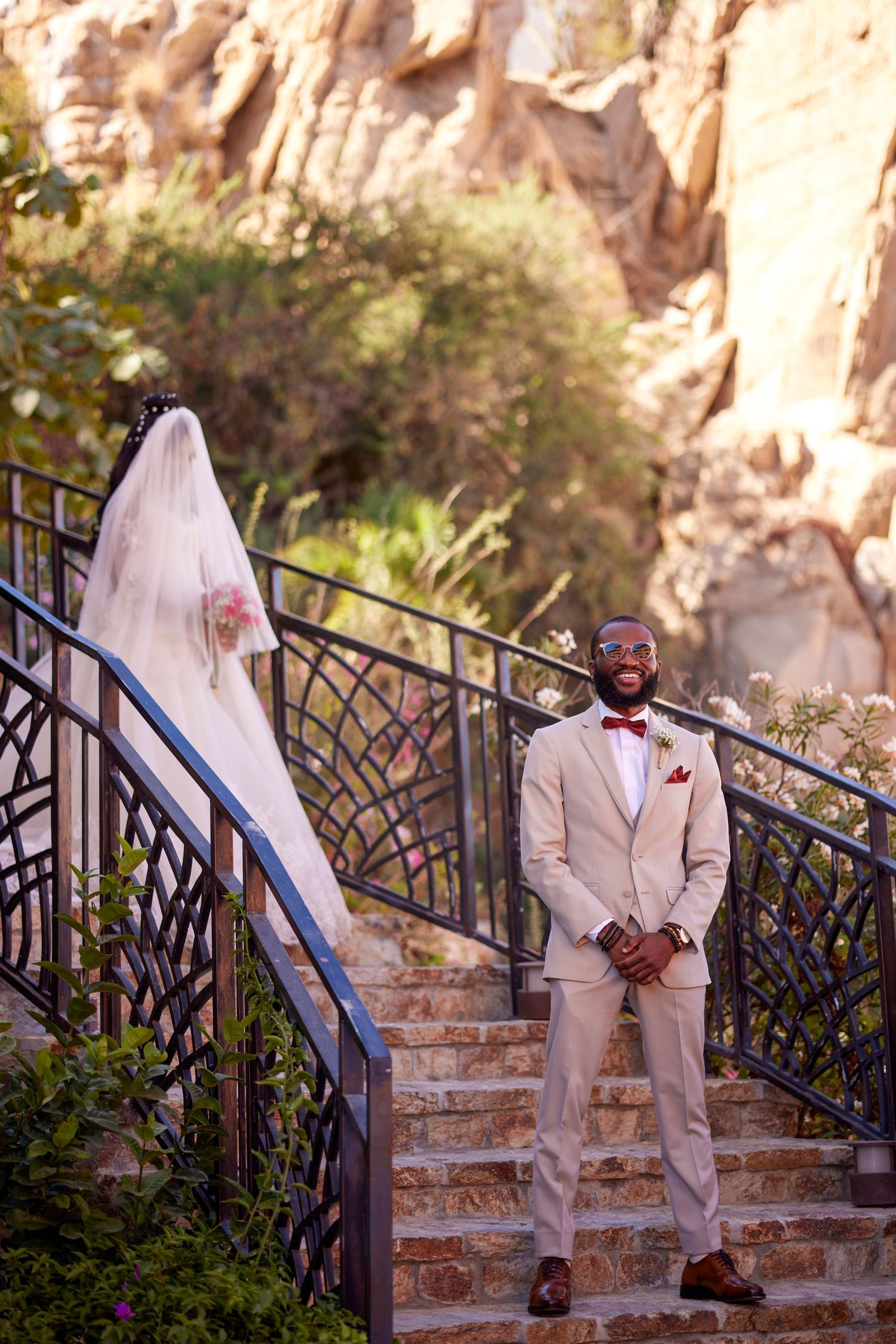 Groom in beige suit and red bow tie stands on stone steps, bride in white veil behind him at outdoor wedding.