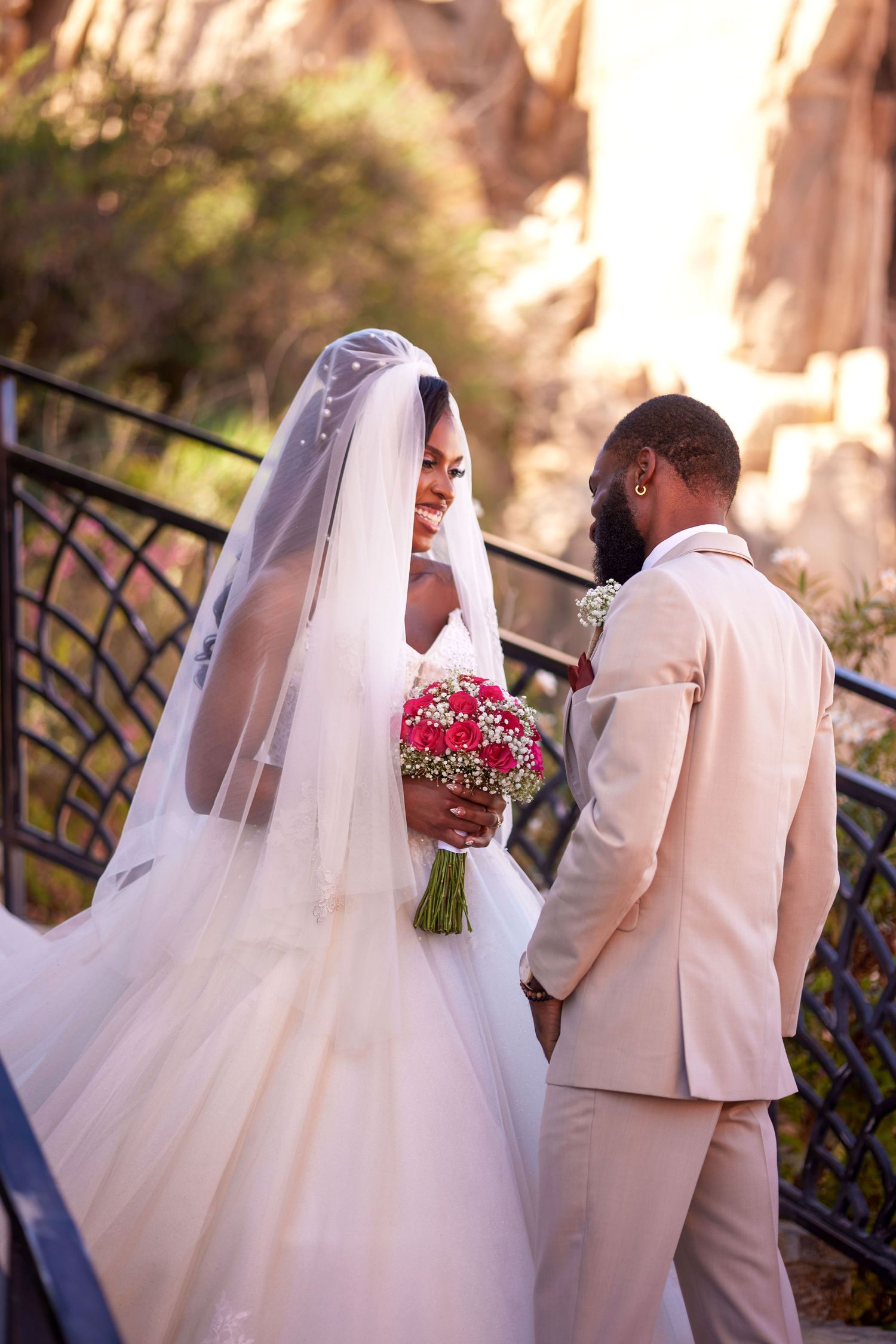 A bride in a white gown and veil holds a red bouquet while a groom in a beige suit gazes at her outdoors.