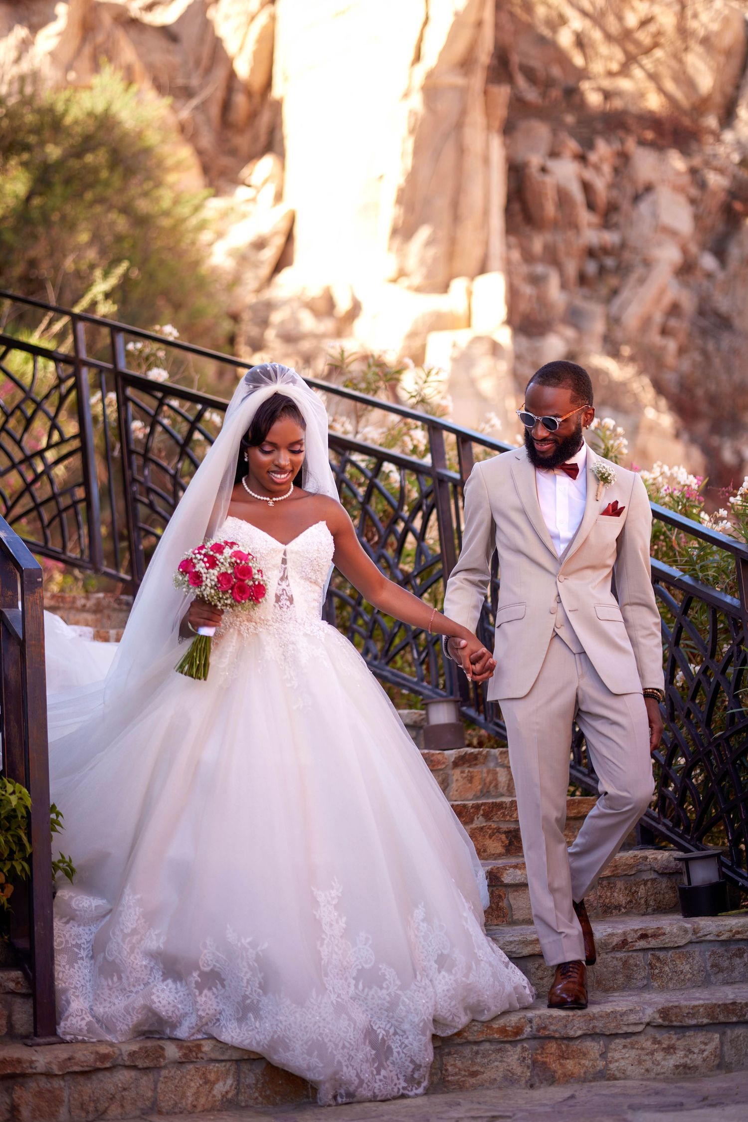 A bride and groom in elegant wedding attire hold hands while descending stone steps outdoors with rocky cliffs behind them.