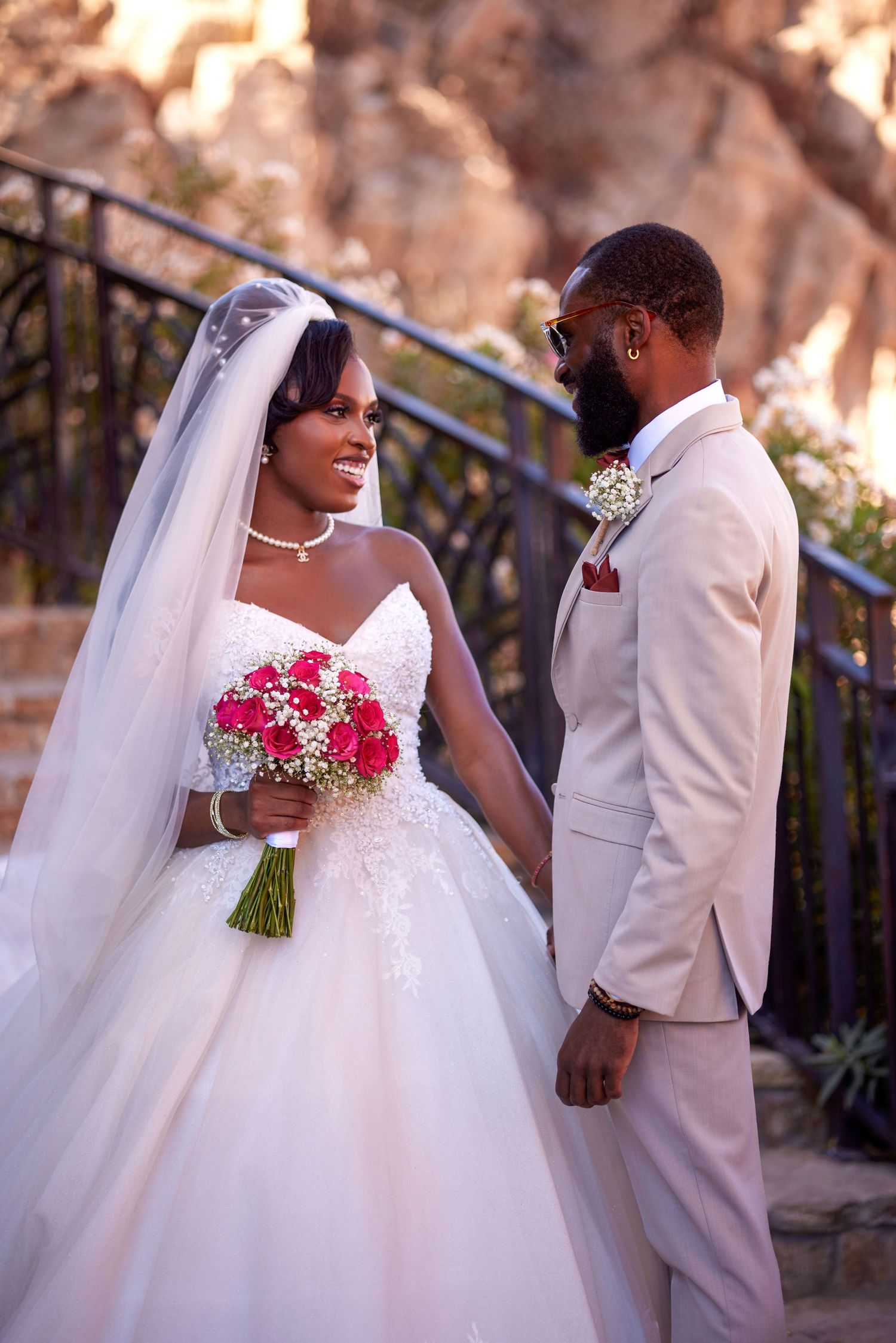 A Black bride in a white ballgown and veil holds red roses while smiling at her groom in a beige suit outdoors.