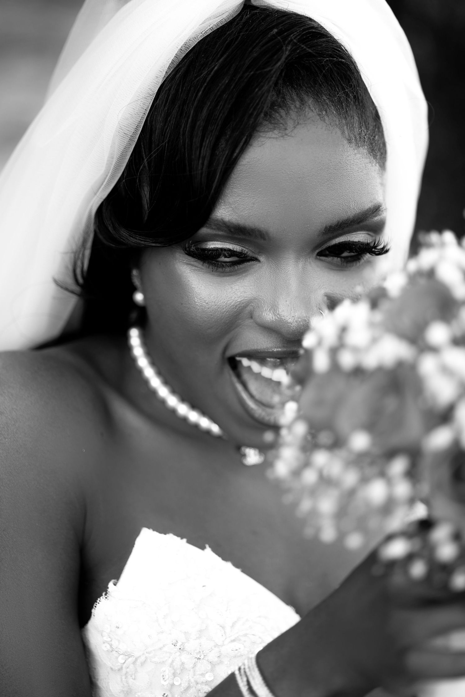 Black and white portrait of a joyful bride in veil and pearl necklace smelling her wedding bouquet.