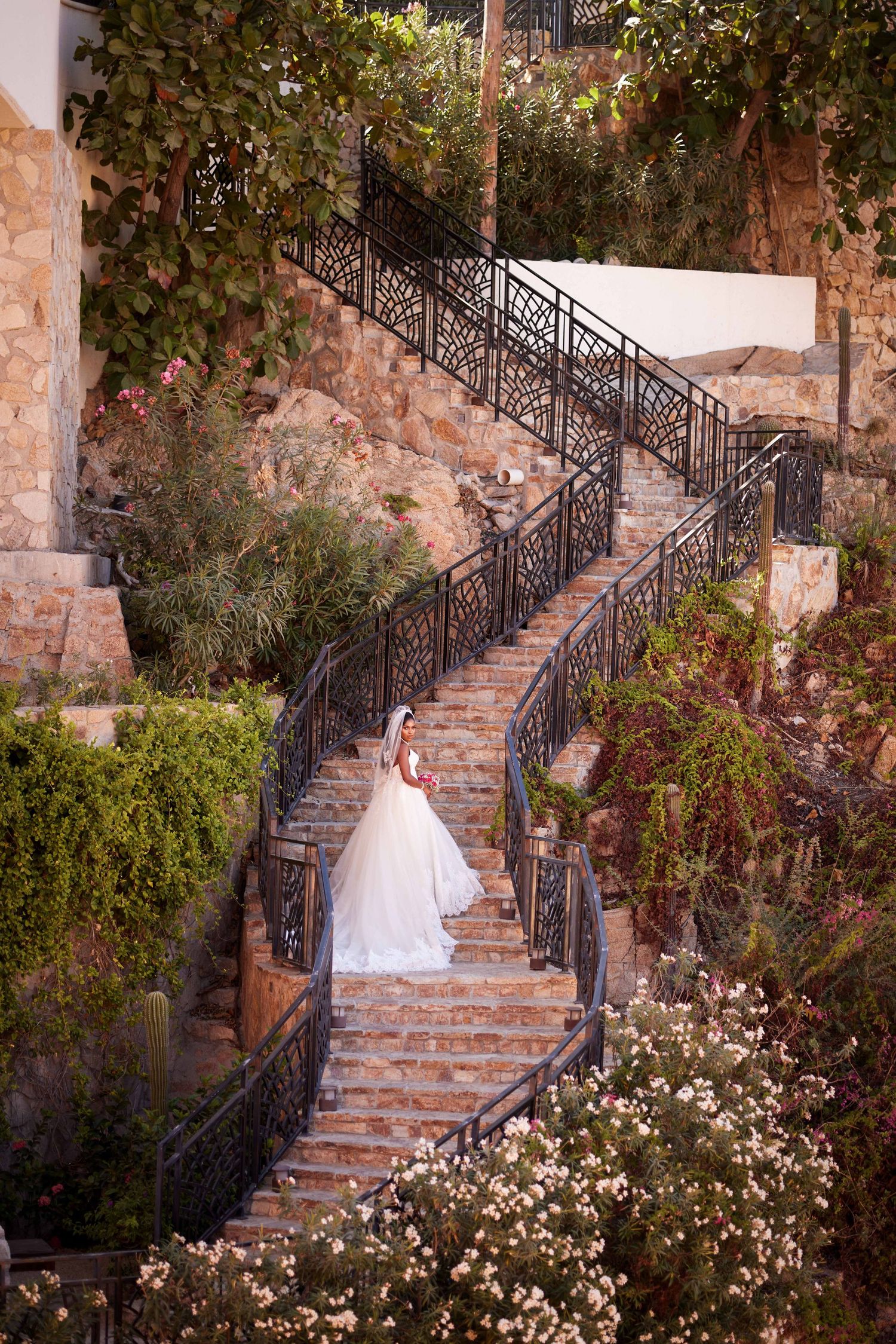 Bride in white gown descending ornate stone staircase surrounded by lush gardens and wrought iron railings.