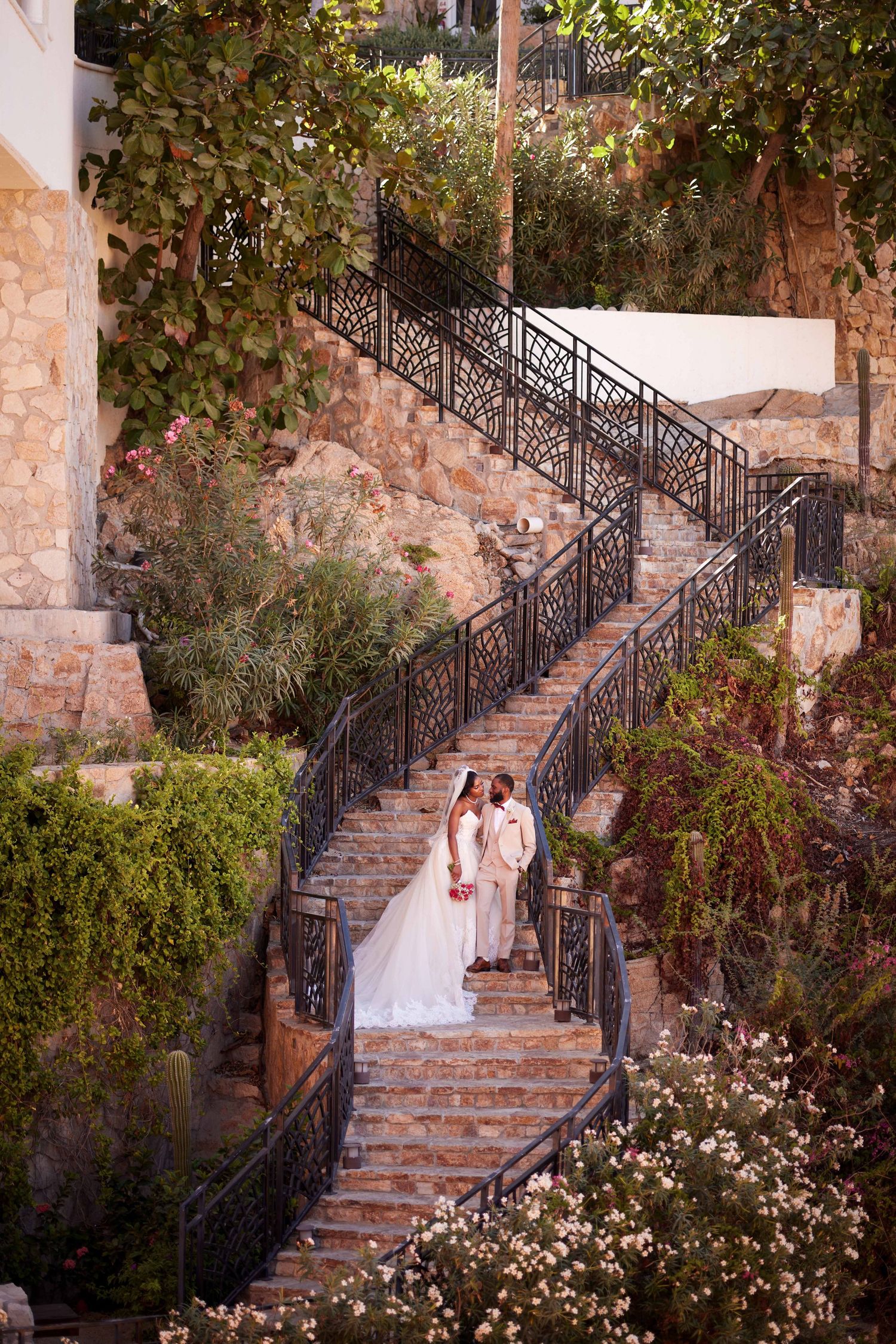 Bride and groom share romantic moment on winding stone staircase surrounded by lush gardens at wedding venue.