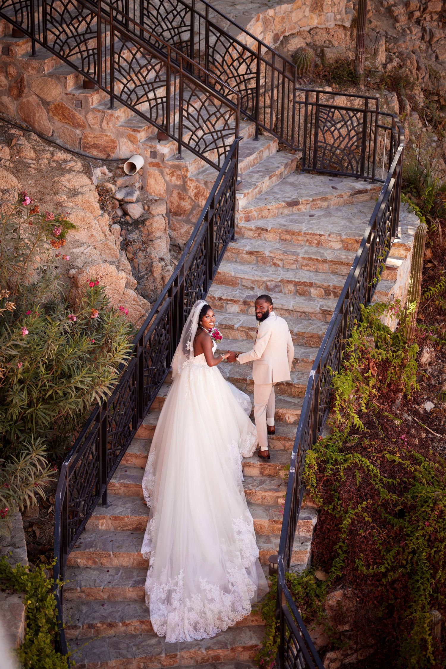 Bride and groom in elegant attire walking down ornate stone stairs surrounded by lush greenery at outdoor wedding venue.