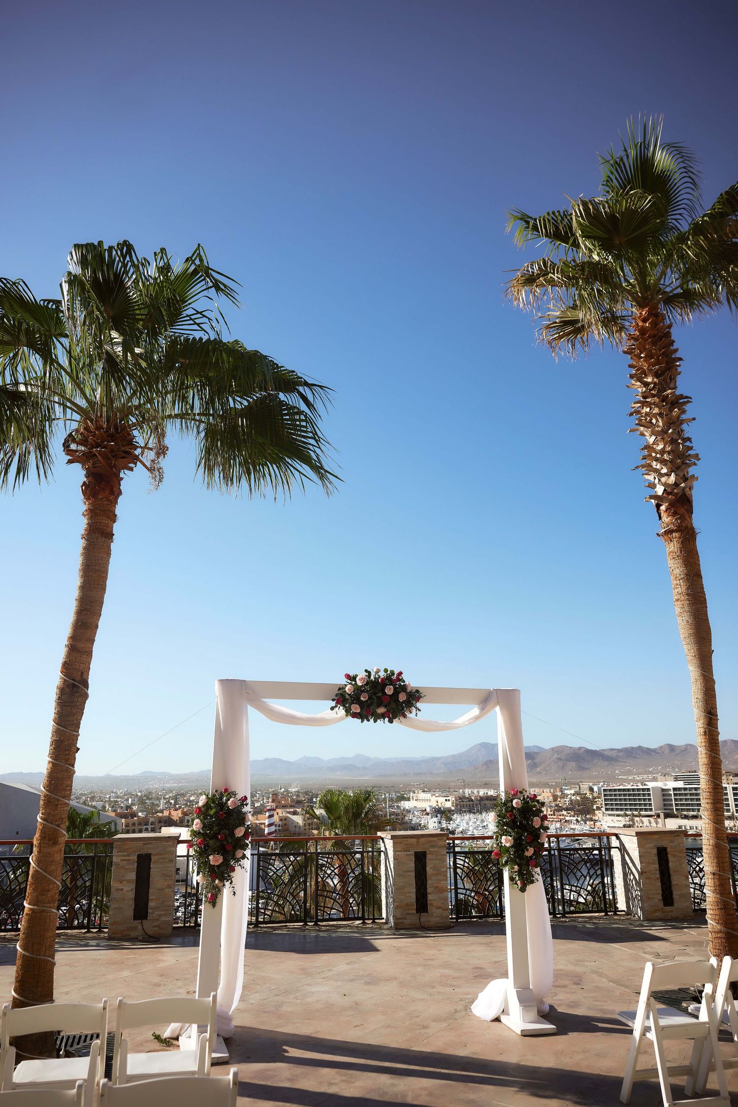 Elegant outdoor wedding arch with white draping and floral decor between palm trees under blue sky.