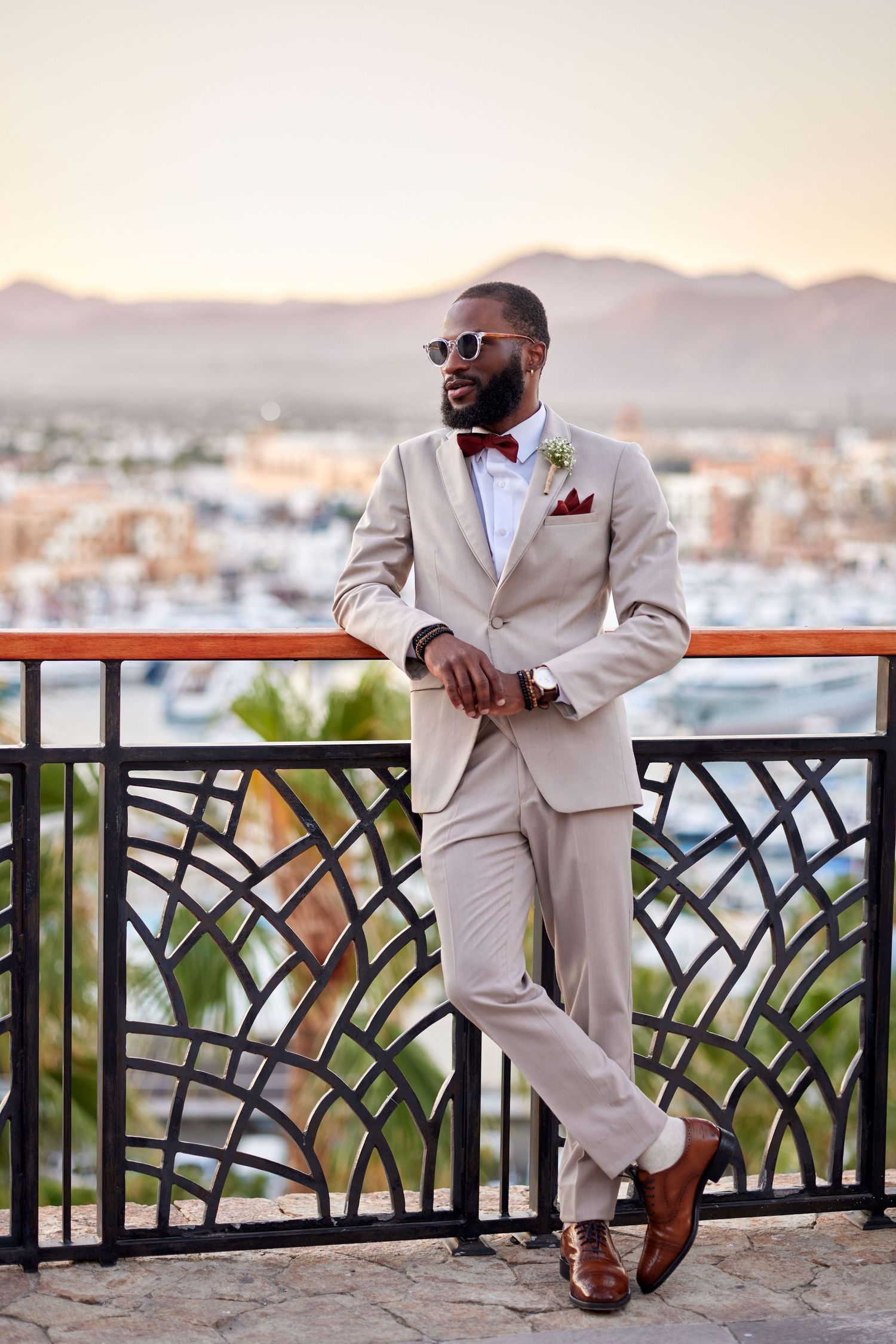 Stylish groom in beige suit with red bow tie posing at railing with scenic city and mountain backdrop.